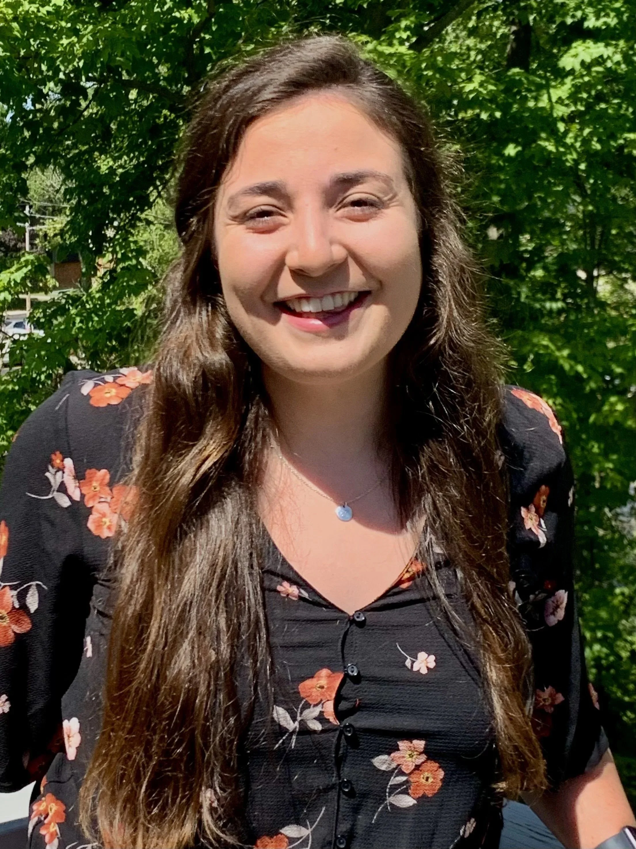 A smiling woman with long brown hair standing outdoors in front of green leafy trees, wearing a black floral blouse and a small pendant necklace.