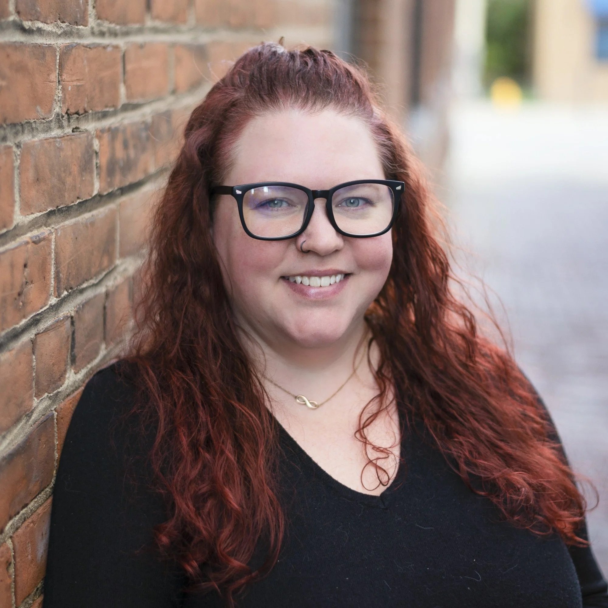 A young woman with curly red hair, glasses, and a nose ring, smiling while leaning against a brick wall outdoors.