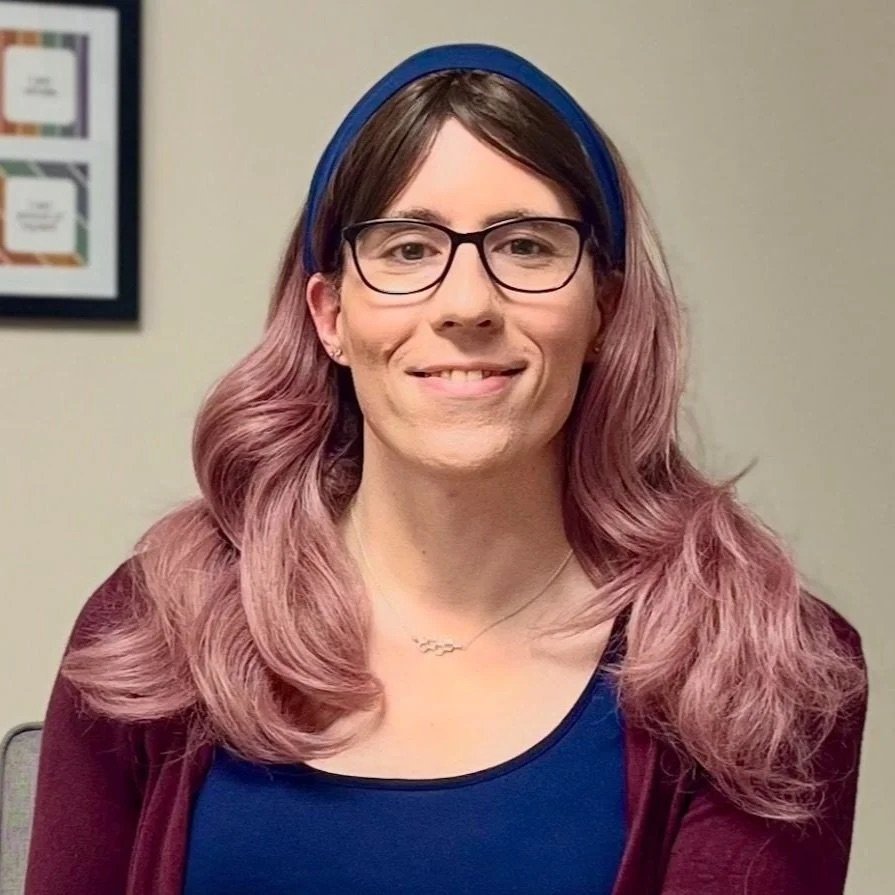 A woman with pink hair, glasses, and a blue headband, smiling in front of a beige wall.