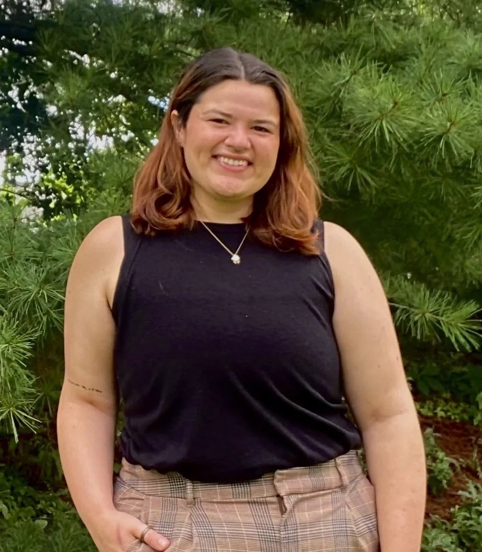 Young woman smiling outdoors in front of green foliage, wearing a black sleeveless top, plaid pants, and a necklace.