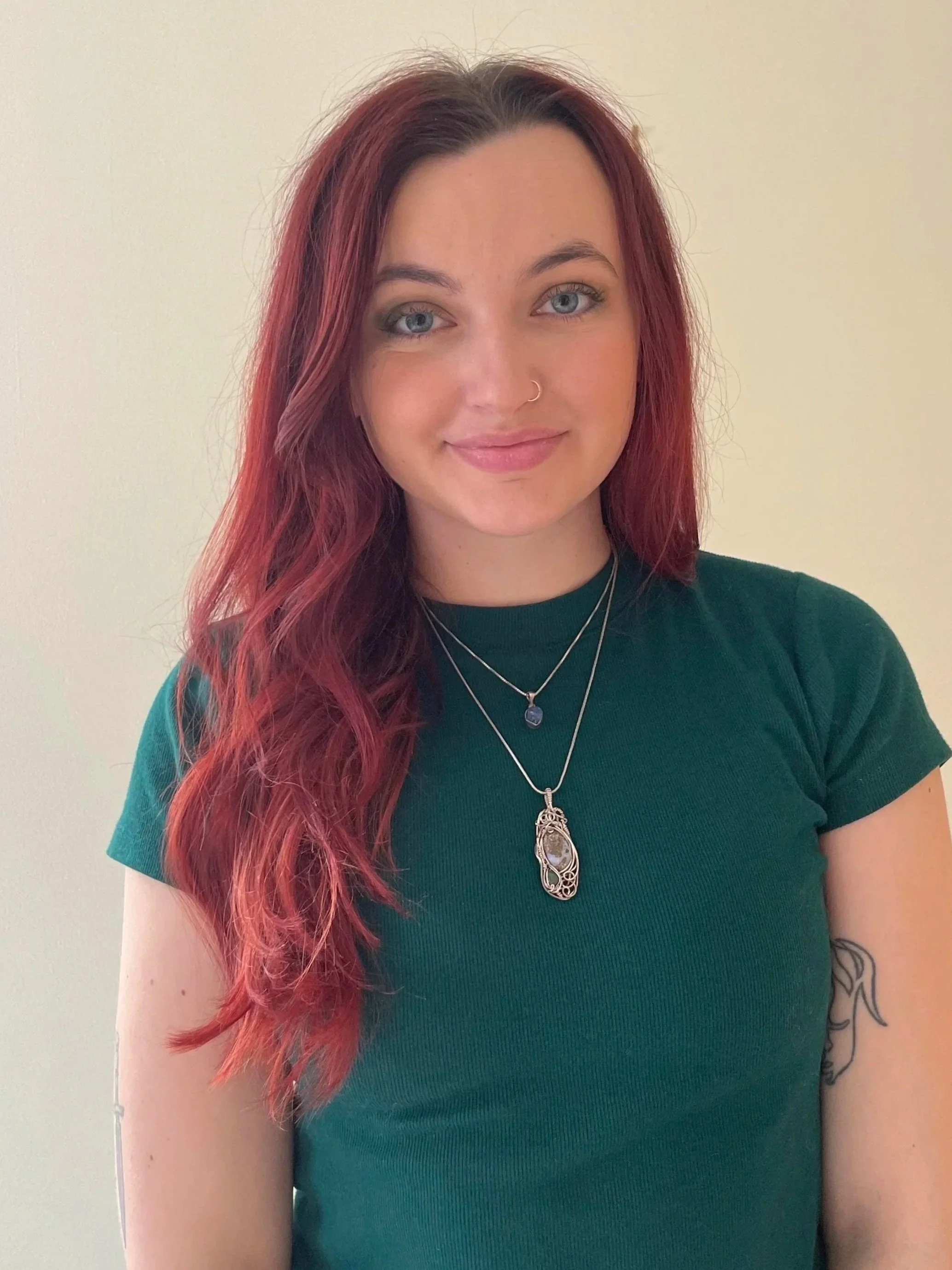 A woman with long, wavy red hair and blue eyes smiling, wearing a dark green t-shirt and layered necklaces, standing against a light-colored background.