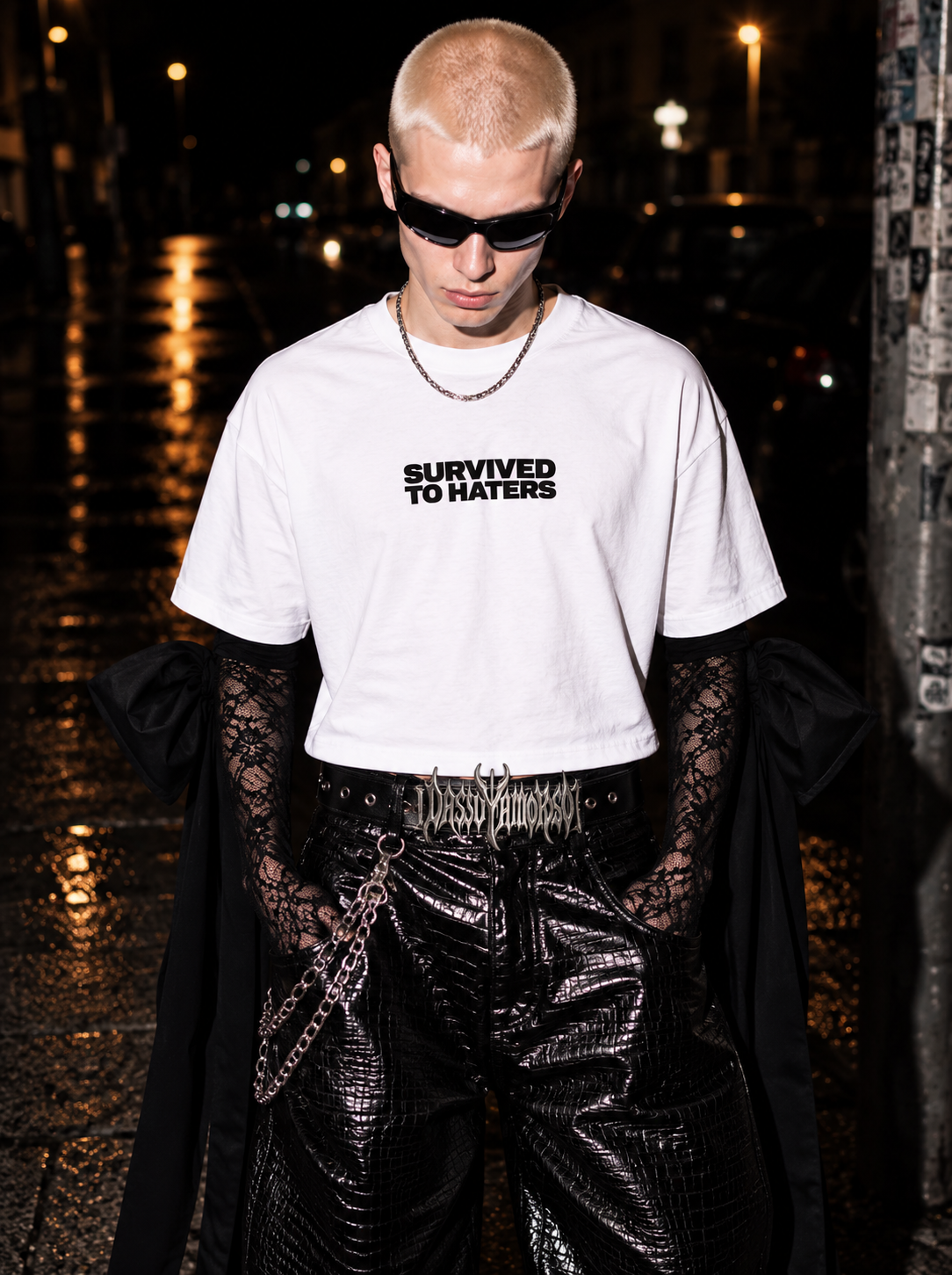 Model wearing a white SURVIVED TO HATERS T-shirt with black lace sleeves and leather trousers on a wet city street at night.