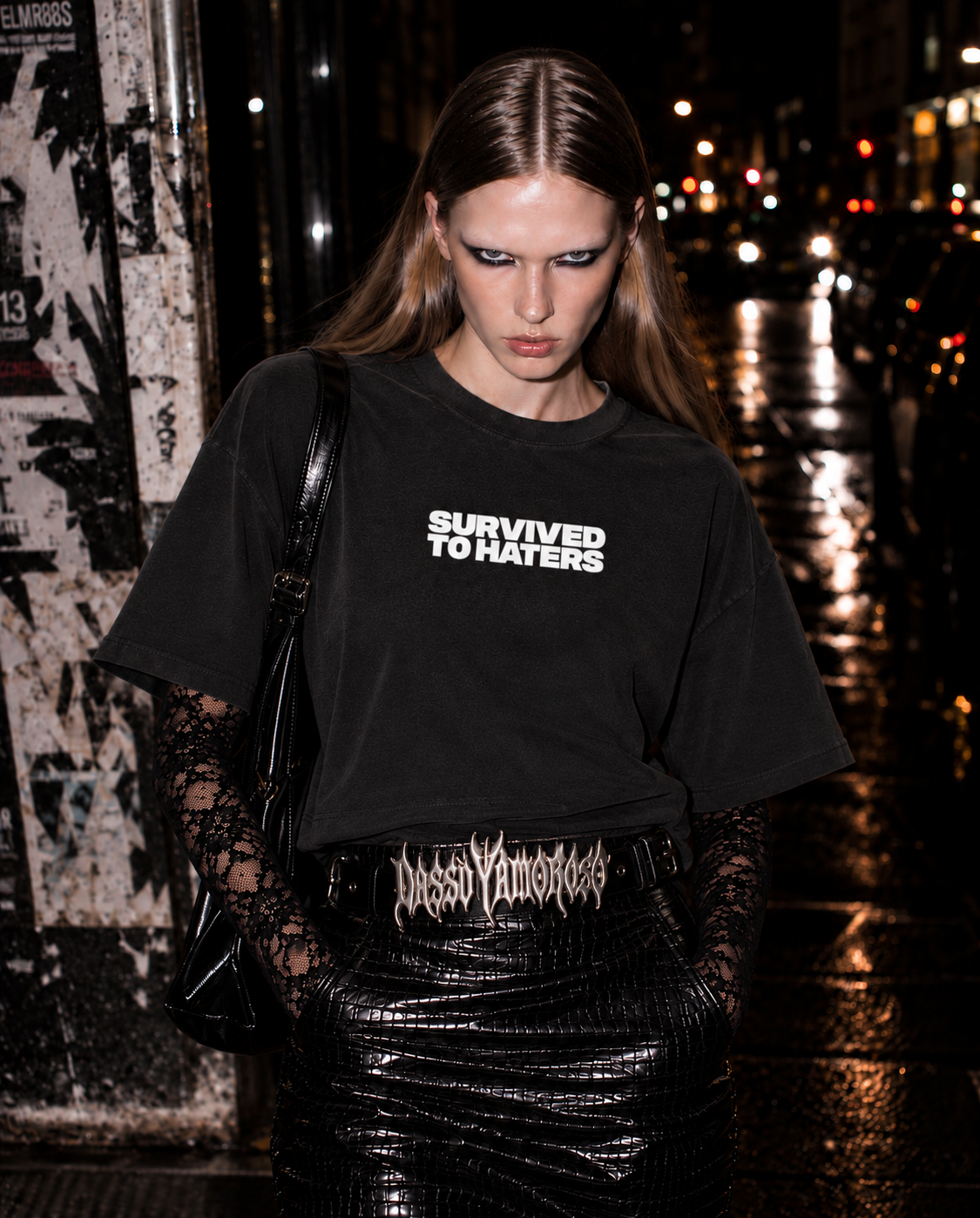 Model wearing a black ICH BIN COUTURE T-shirt with lace sleeves and a gothic belt on a wet city street at night.