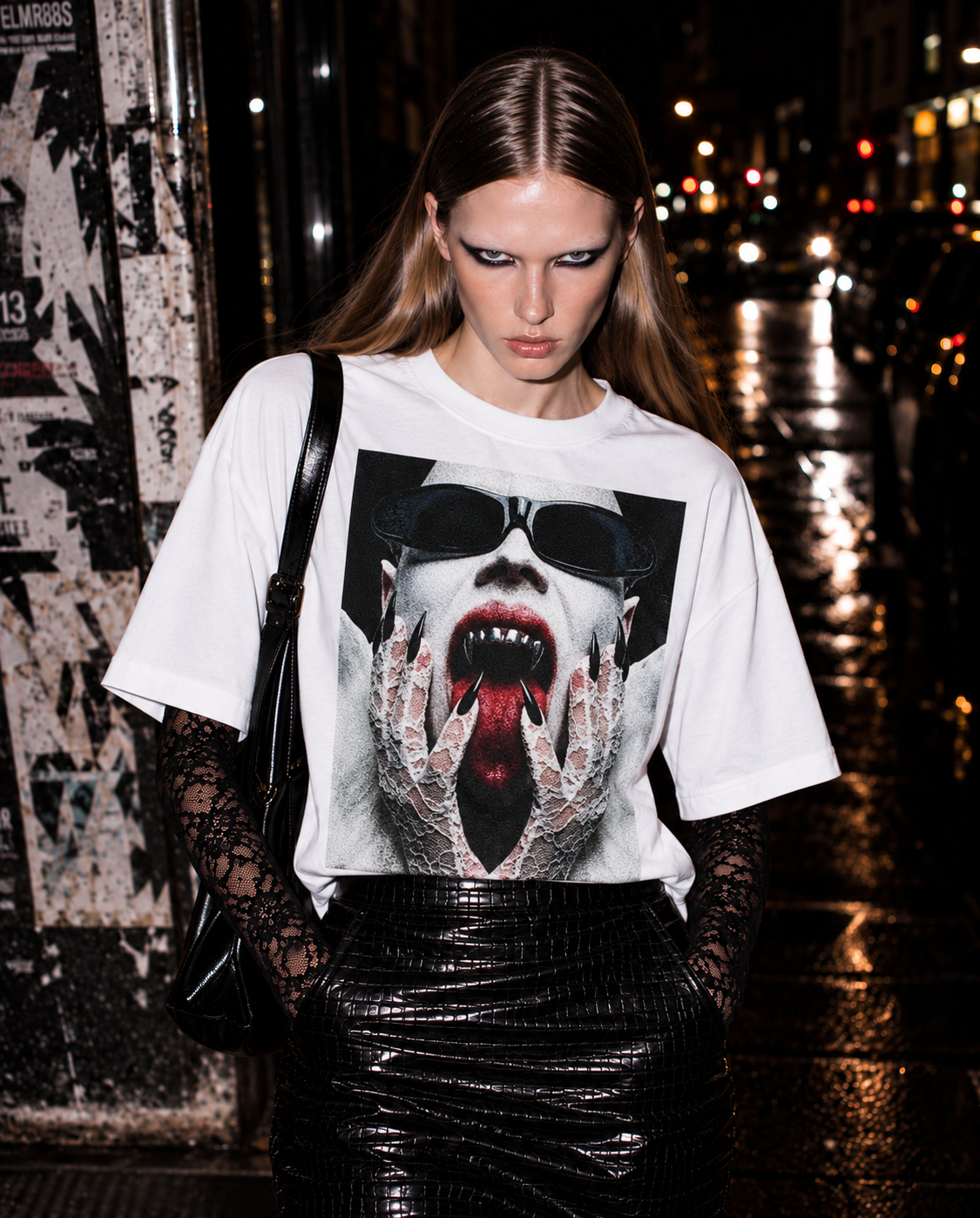 Model wearing a white graphic T-shirt with a vampire-inspired design layered over black lace on a city street at night.