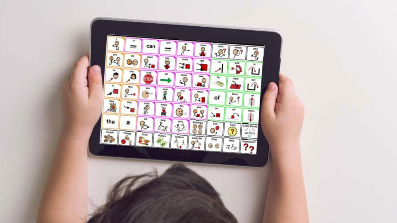 Child holding a tablet displaying a colorful picture communication board with various words and pictures.