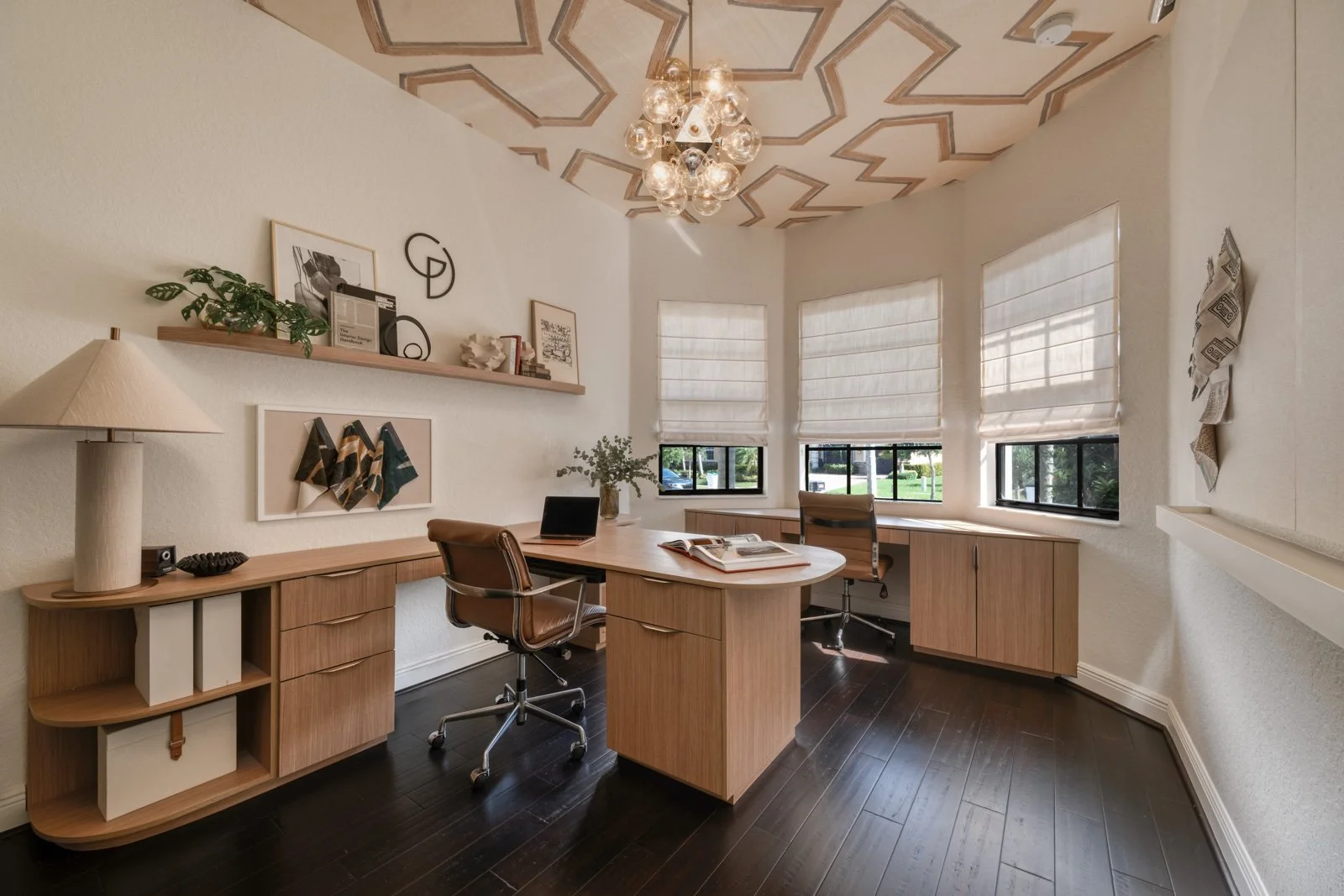 Wide-angle view of a Parkland FL home office featuring custom wood desk, built-in shelving, textured table lamp, floating wall shelf with decor, and roman shade windows designed by a South Florida interior designer