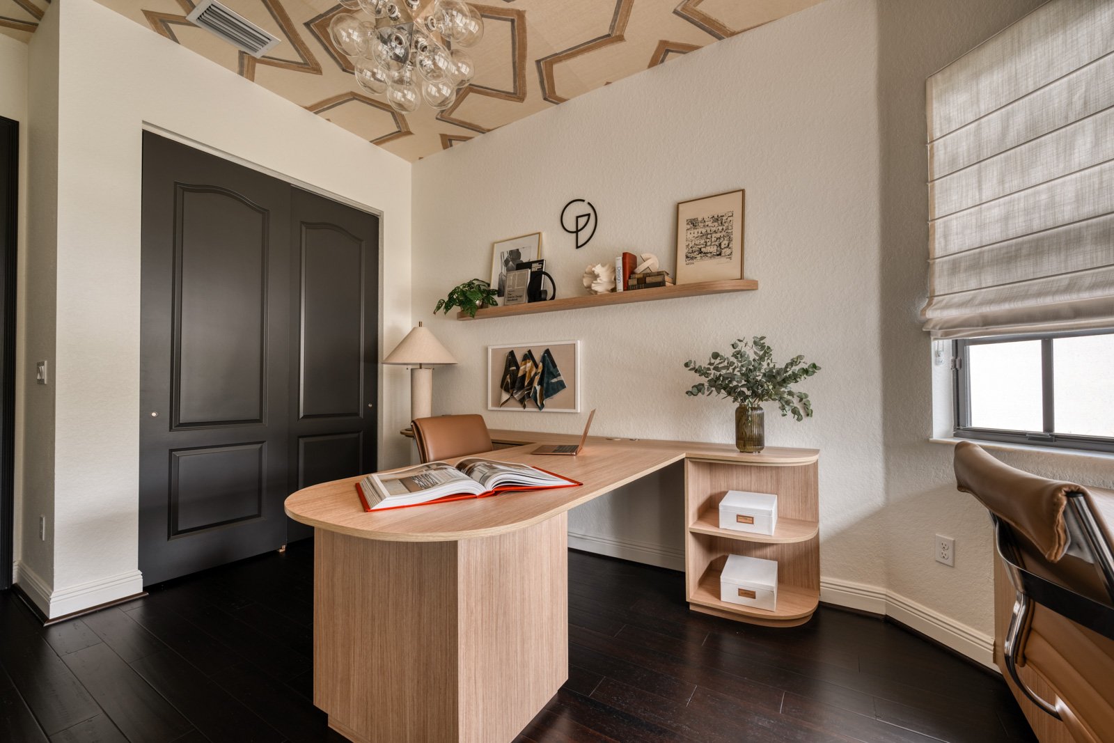 arkland FL home office entry view with dark cupboard doors, custom light wood L-shaped desk, floating shelf with curated objects, and a sculptural bubble chandelier against a geometric wallpaper ceiling