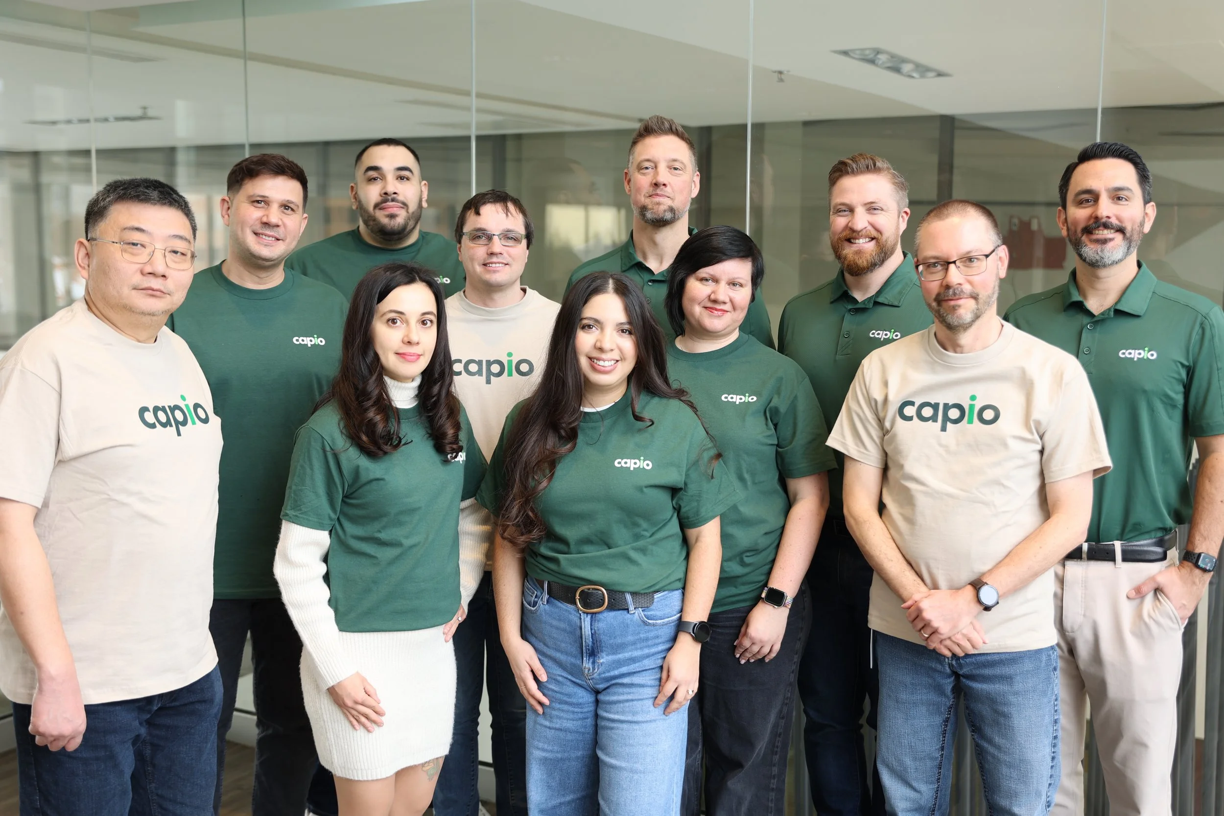 Group of ten diverse people standing together indoors, wearing green and beige shirts with the word 'capio' printed on them, smiling and looking at the camera.