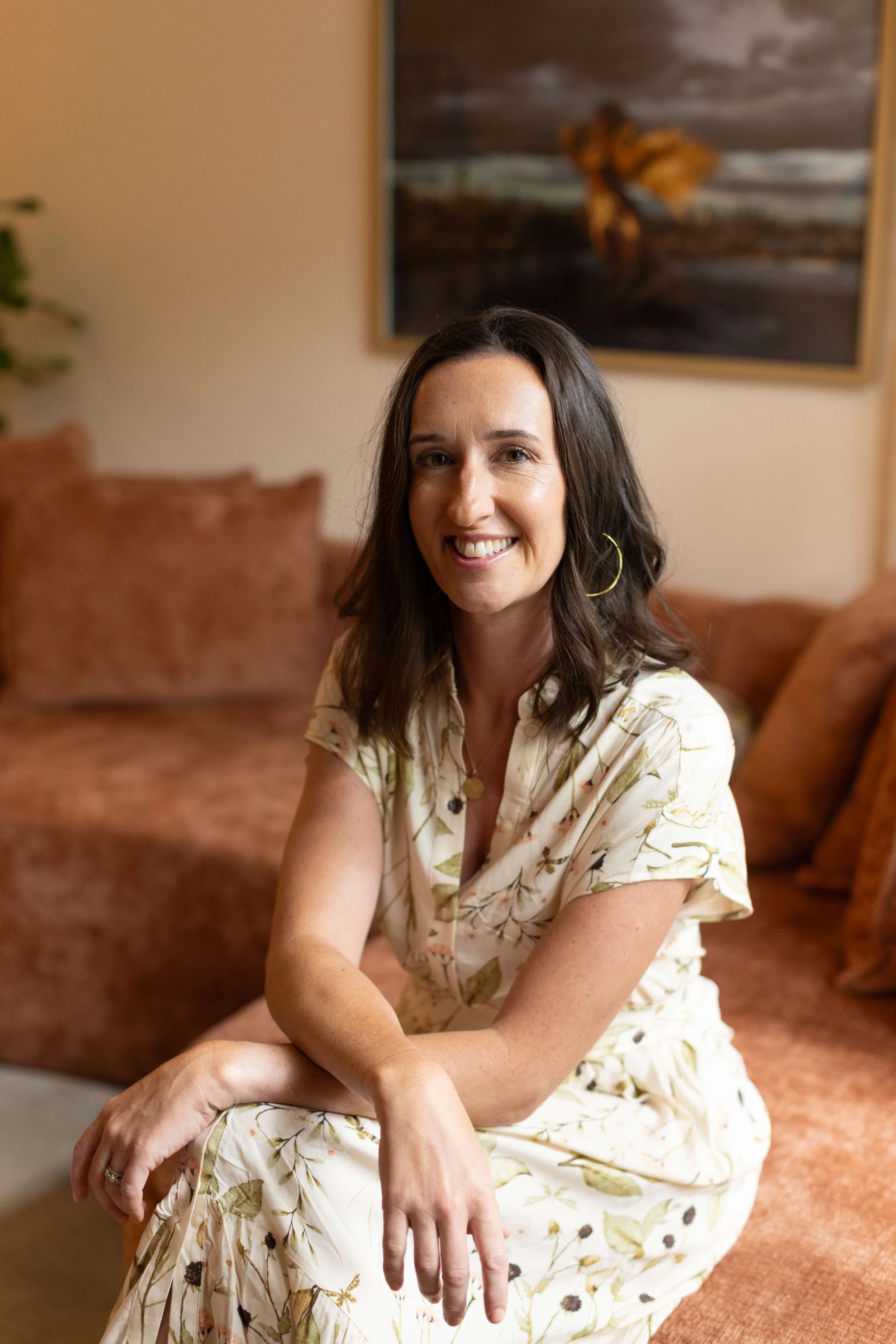 Sarah Meehan, LCSW, is sitting on a sofa, smiling at the camera. Sarah specializes in general psychotherapy, perinatal/postpartum therapy and support groups in NYC.