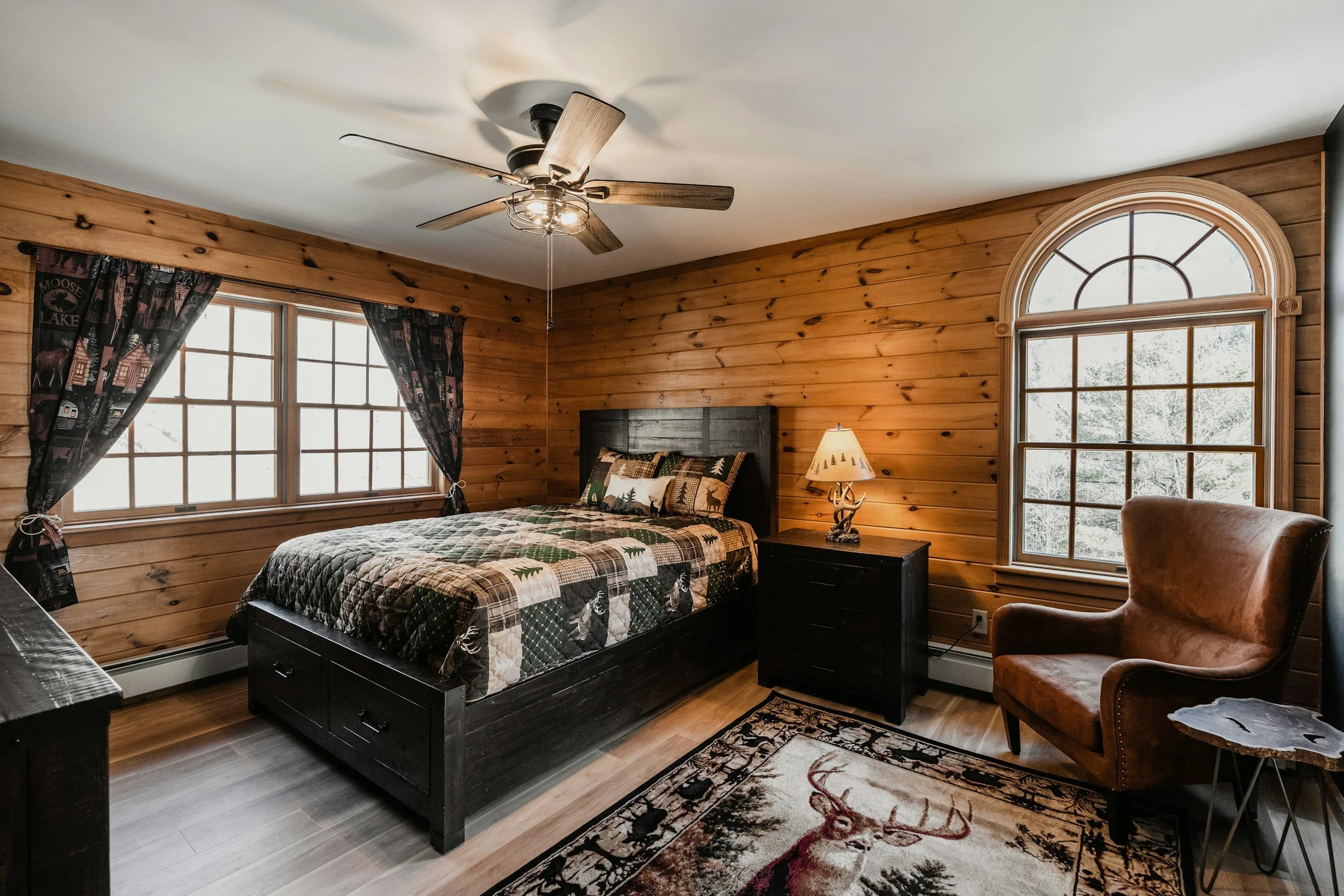 A cozy bedroom with wooden paneled walls, a large bed with a quilted bedspread, a nightstand with a lamp, a leather armchair, and windows with mountain views.