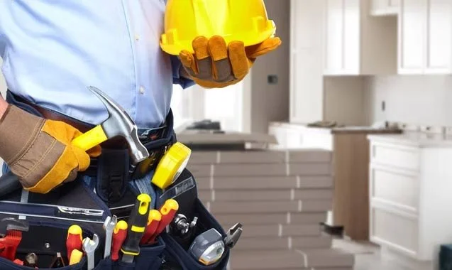 Close-up of a contractor with yellow gloves holding a yellow hard hat, dressed in a tool belt with various tools, inside a kitchen under construction.