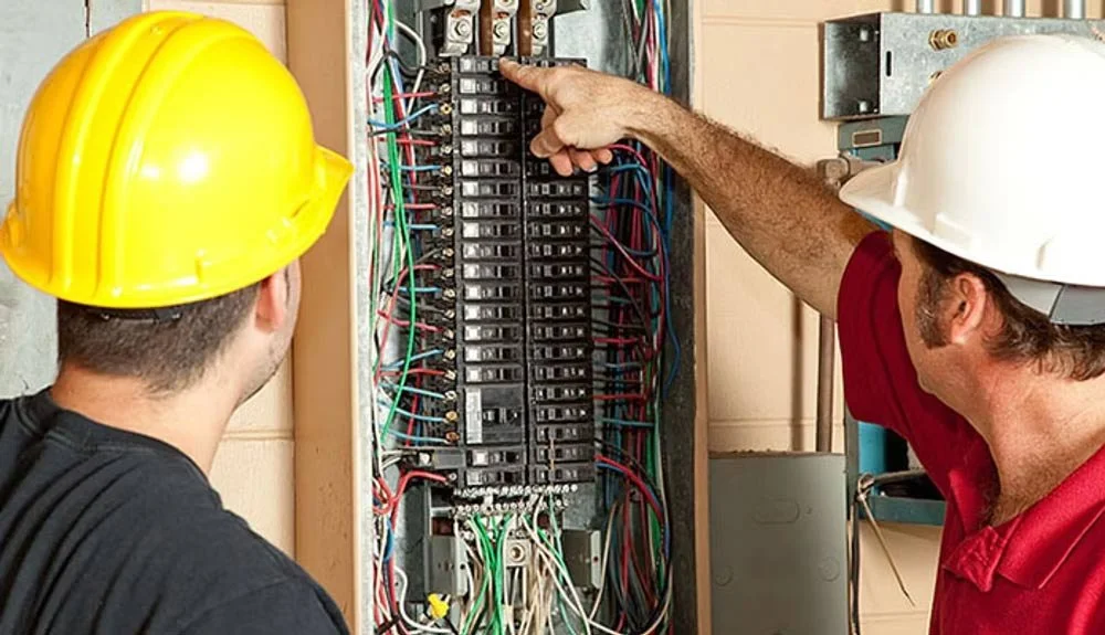 Two electricians wearing safety helmets working on an electrical panel with multiple wires and circuit breakers.