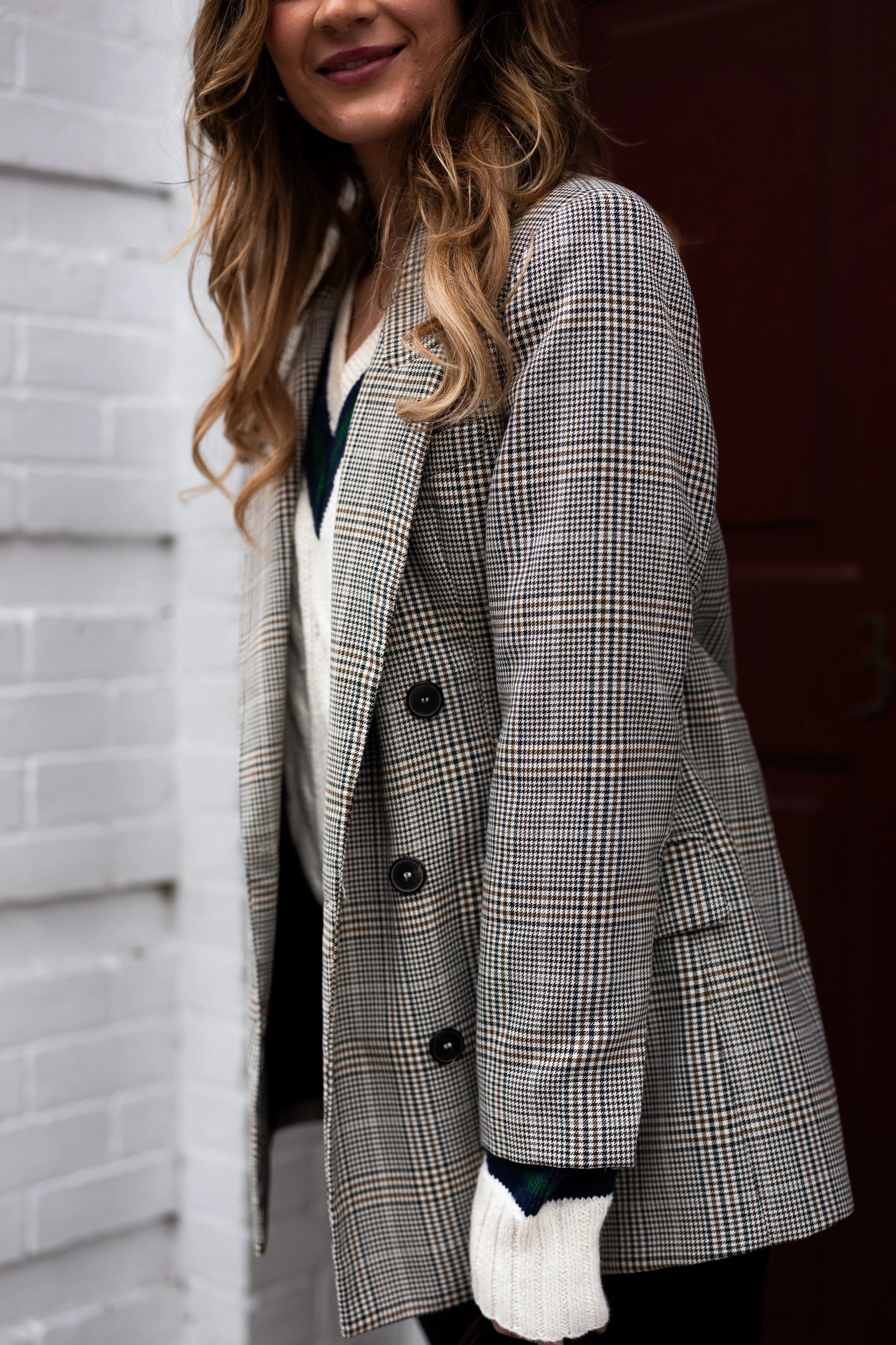 A woman with long wavy hair wearing a plaid blazer over a sweater, standing in front of a white brick wall and a red door, smiling.
