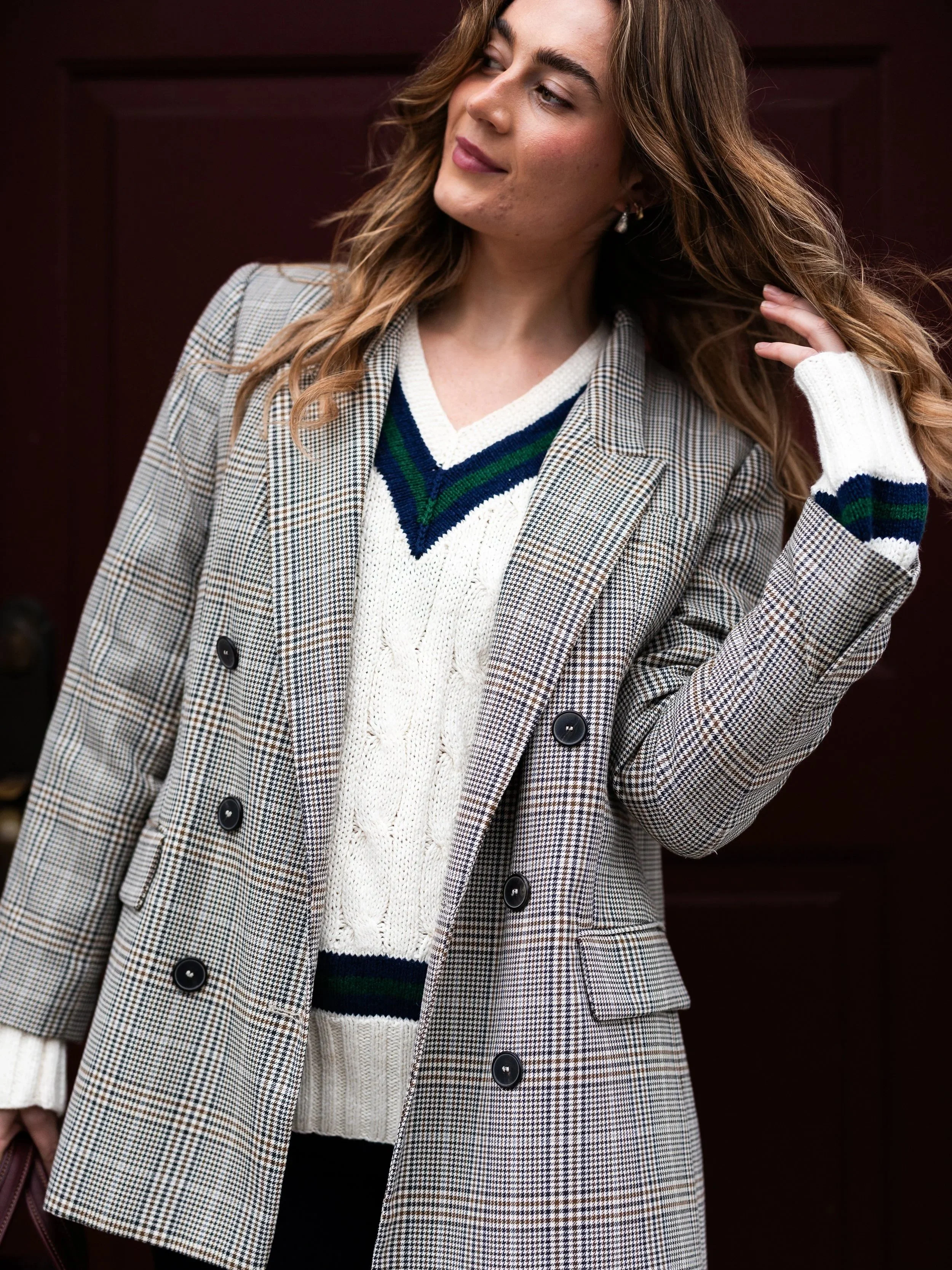A woman with long, wavy brown hair wearing a plaid double-breasted coat over a white sweater with green and navy stripes. She is touching her hair with her right hand and standing in front of a dark brown background.