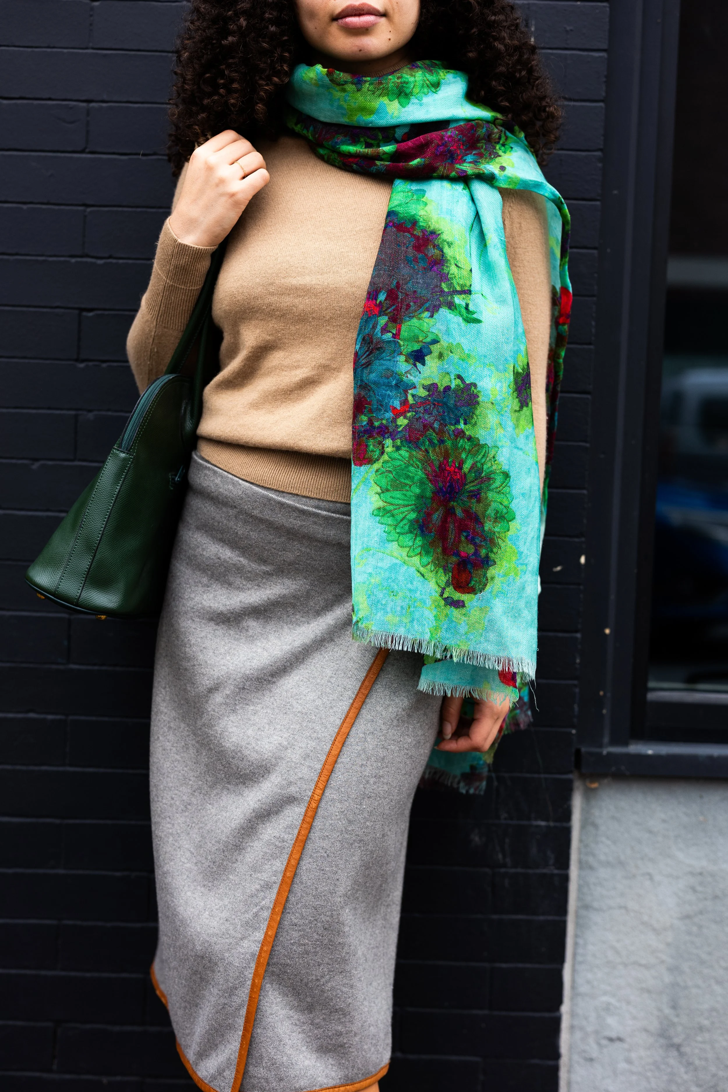 A woman standing against a black brick wall, wearing a beige sweater, a gray skirt with an orange stripe, and a colorful scarf, carrying a green handbag.