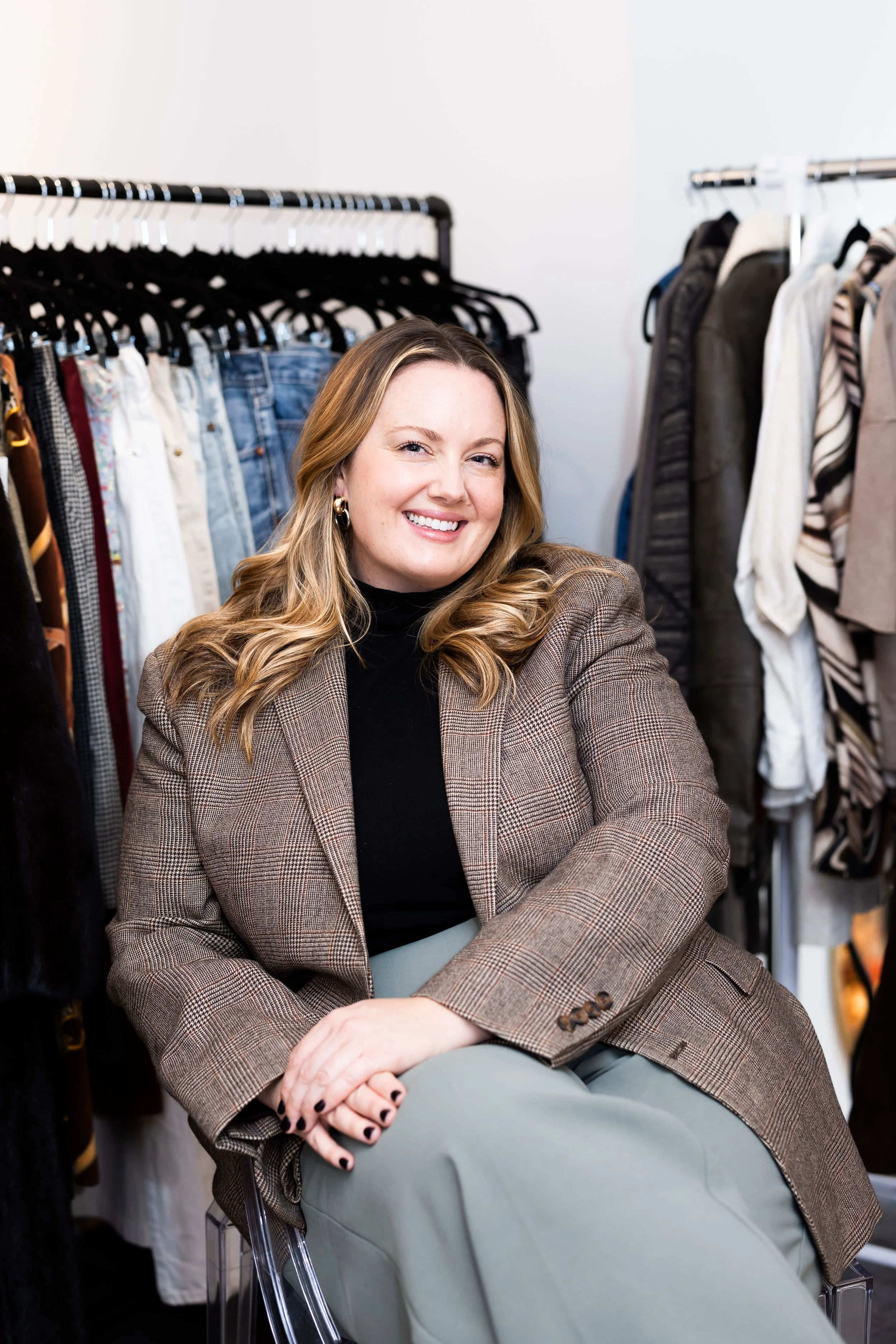 A woman with blonde hair, wearing a checkered blazer and black turtleneck, sitting and smiling in a clothing store with racks of jeans and jackets in the background.