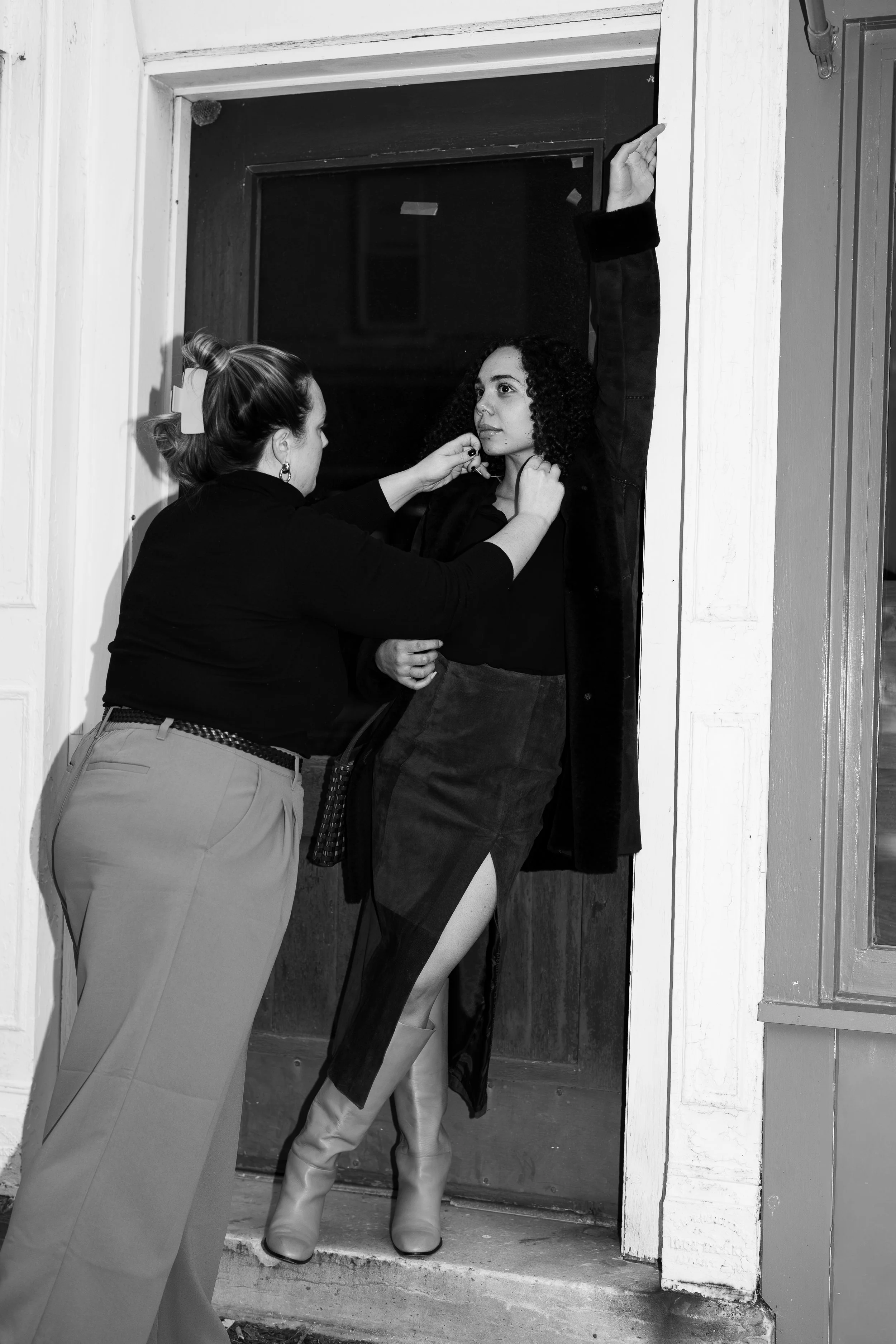 A woman is applying makeup on a young woman who is standing against a doorframe, with one arm raised and the other hand resting on her neck.