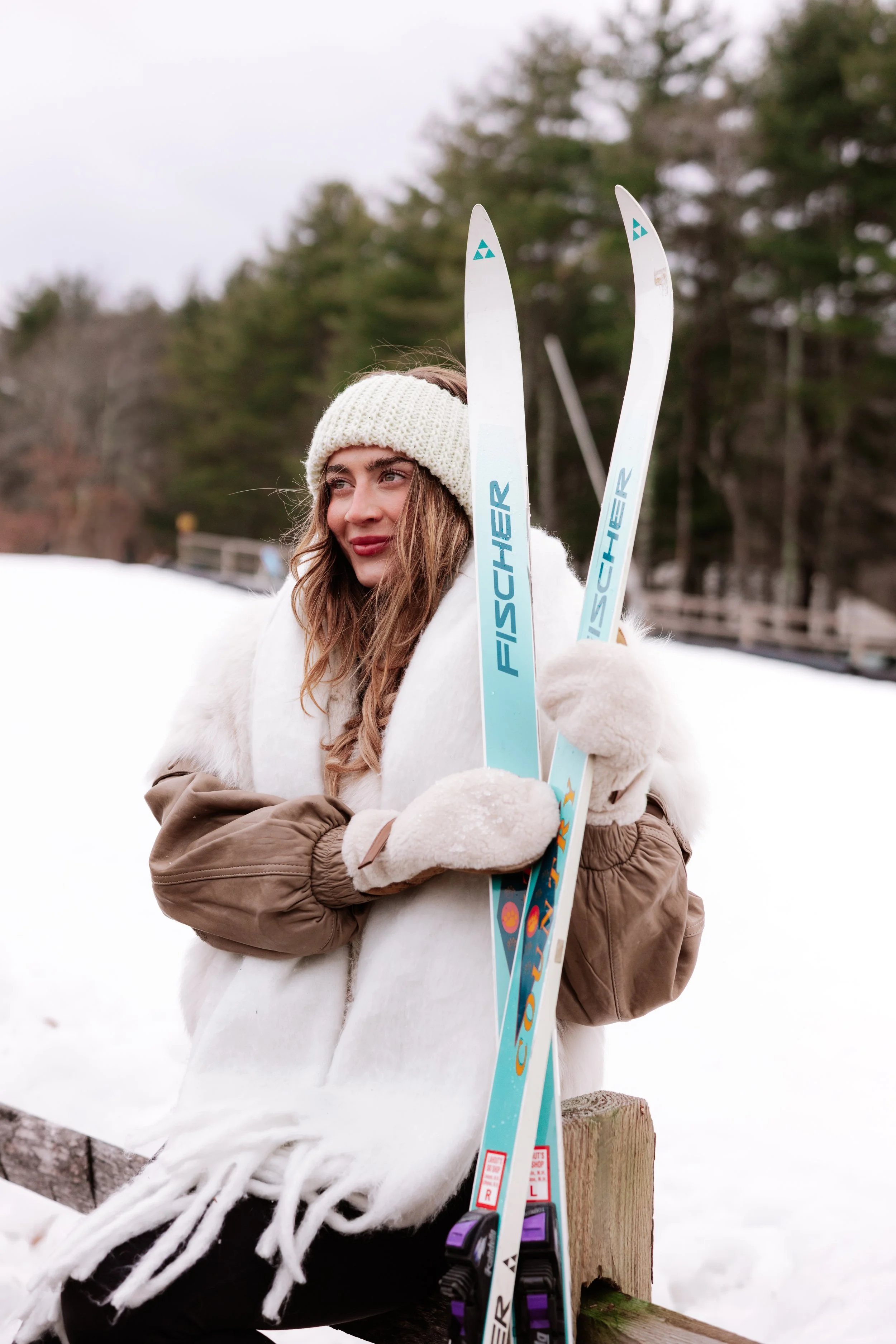 Young woman in winter clothing holding skis outdoors in snow-covered area with trees in background.