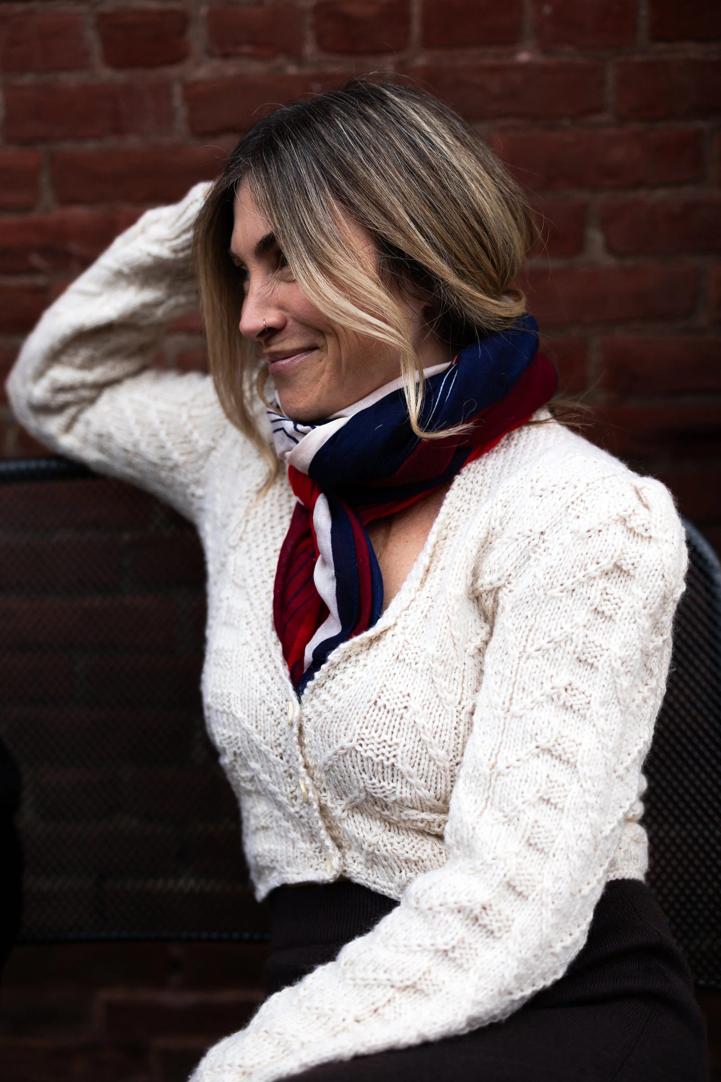 A woman with shoulder-length blonde hair, wearing a white knit sweater and a red, white, and blue scarf, sitting against a brick wall, with a smile and her eyes closed, resting her hand on her head.