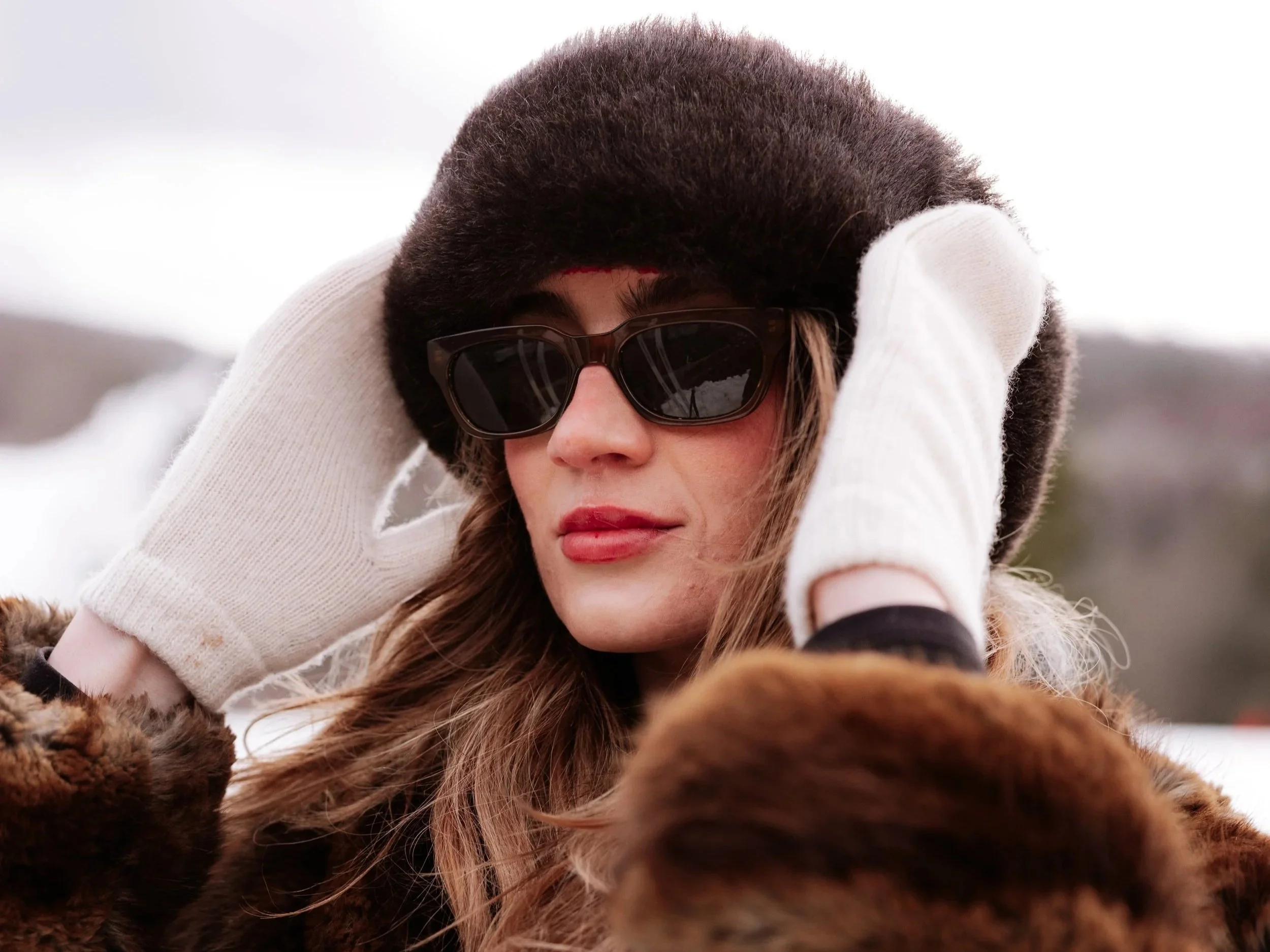 Woman wearing a fur hat, sunglasses, white gloves, and a fur coat outdoors in winter.