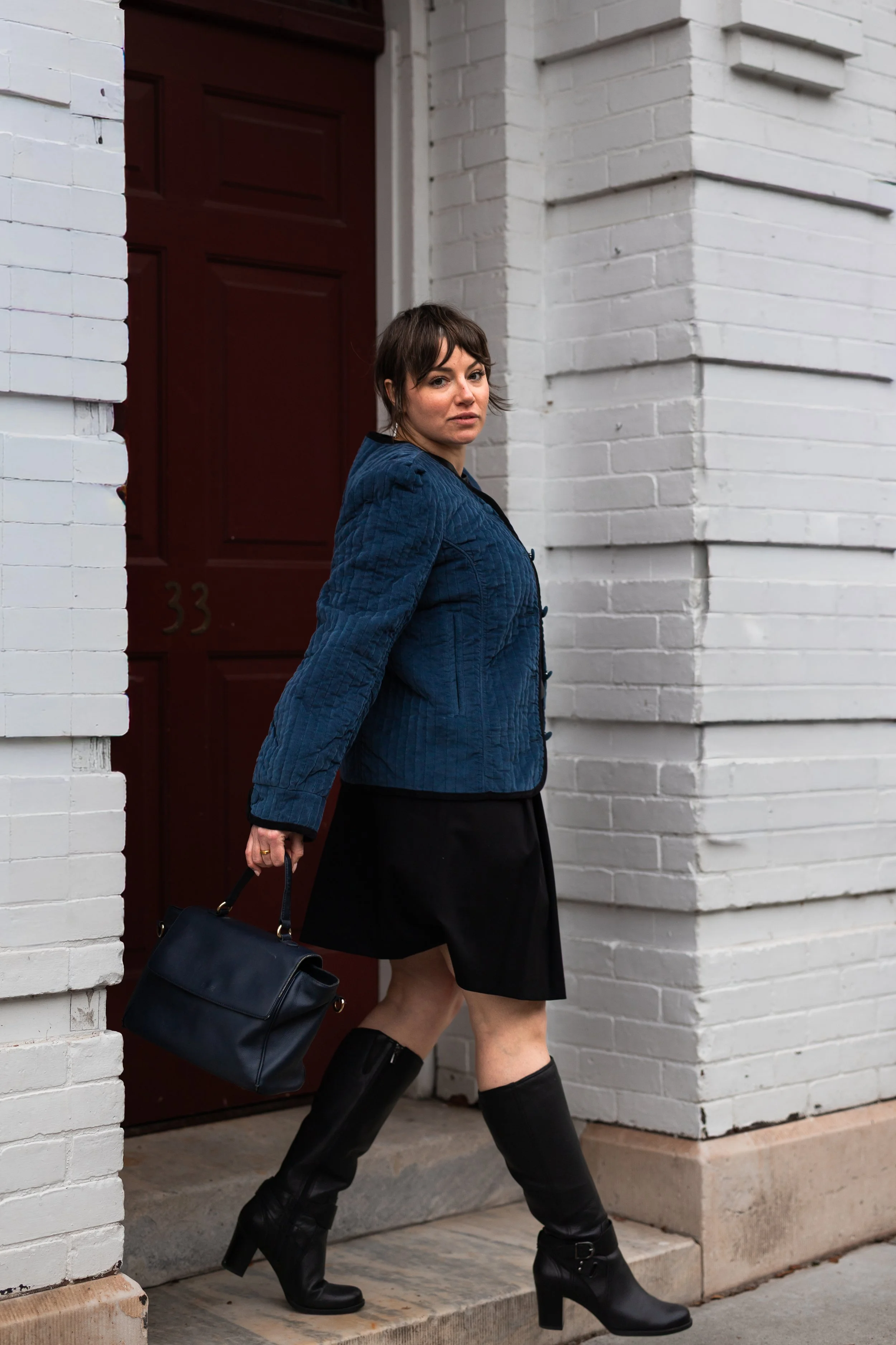 A woman with short brown hair wearing a blue jacket, black skirt, and black knee-high boots carrying a black handbag and standing near a white brick wall with a red door numbered 33.