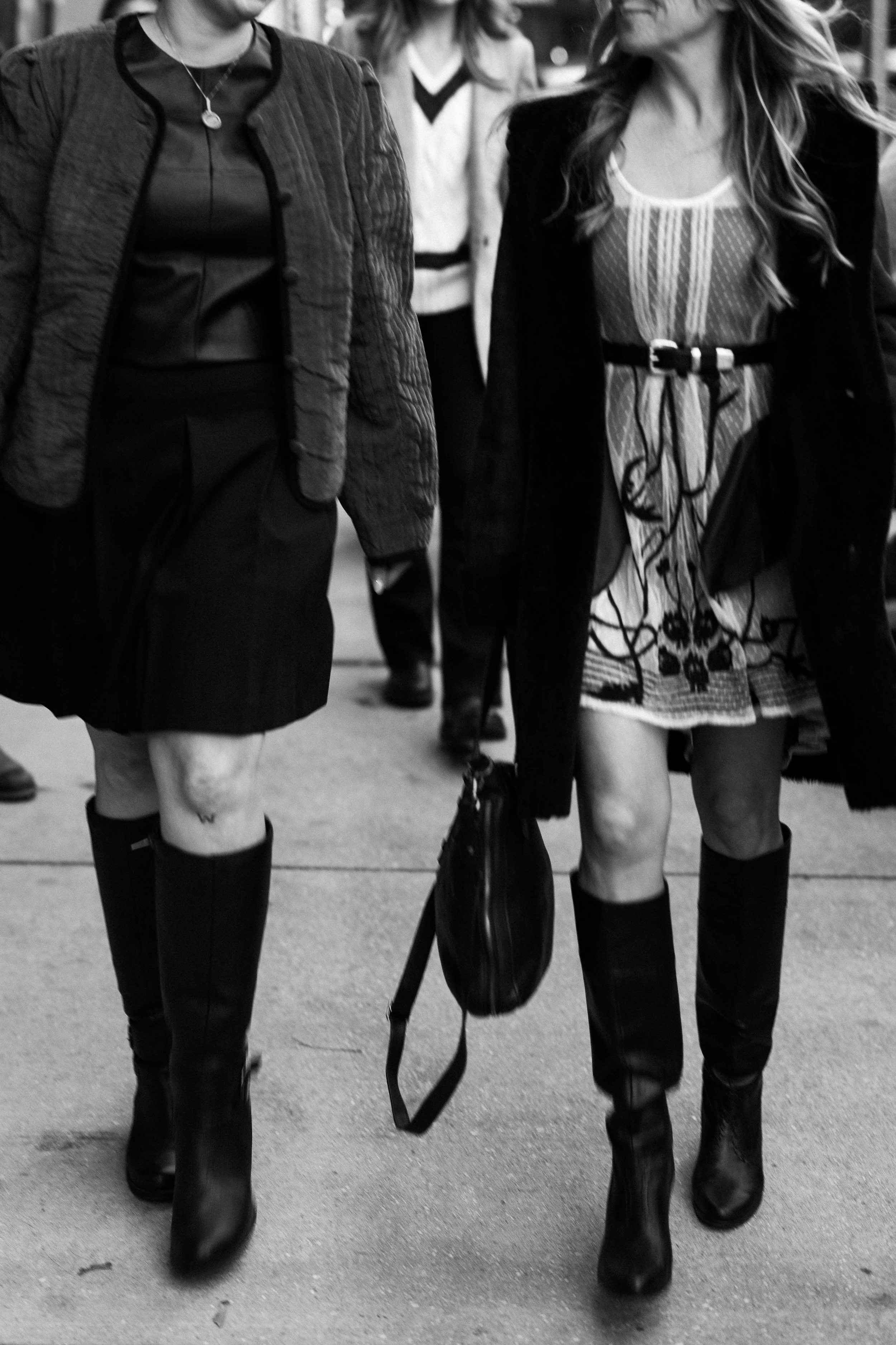 Two women walking in stylish outfits, black knee-high boots, and carrying a shoulder bag, seen from the waist down in black and white photograph.