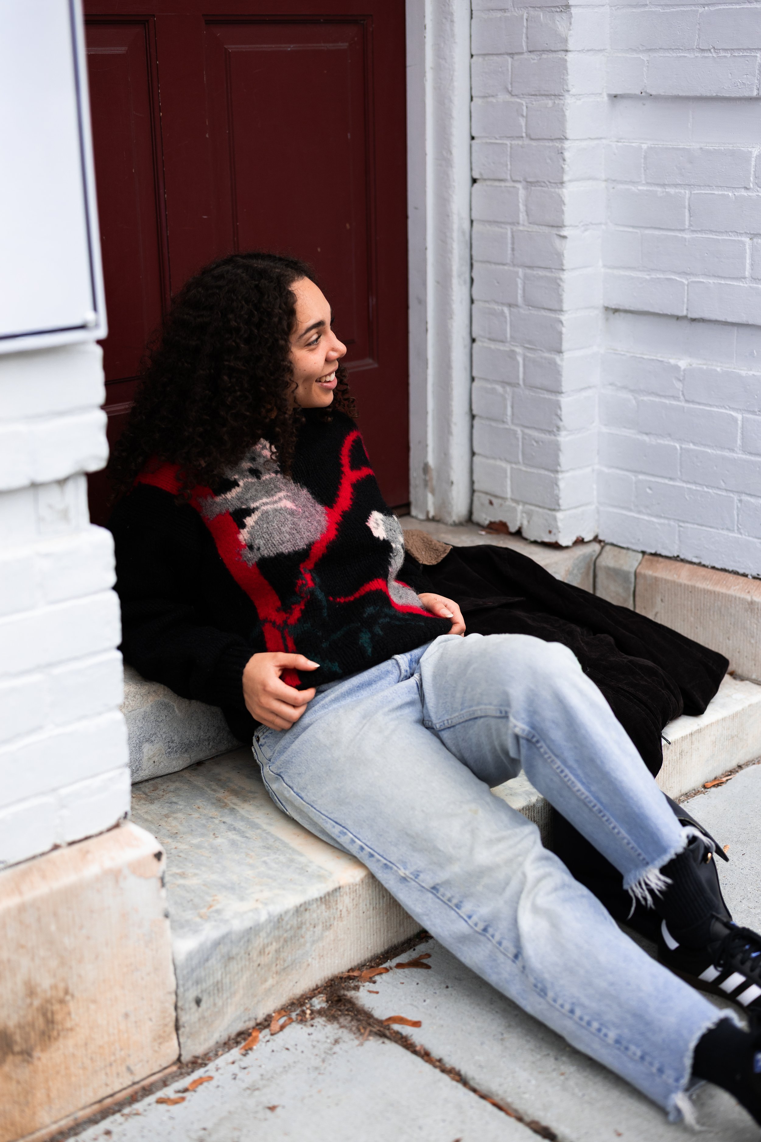 A young woman sitting on a concrete step outside a building with a white painted brick wall and a red door, smiling and leaning back with hands on her stomach, wearing a colorful sweater, light blue ripped jeans, and black sneakers.