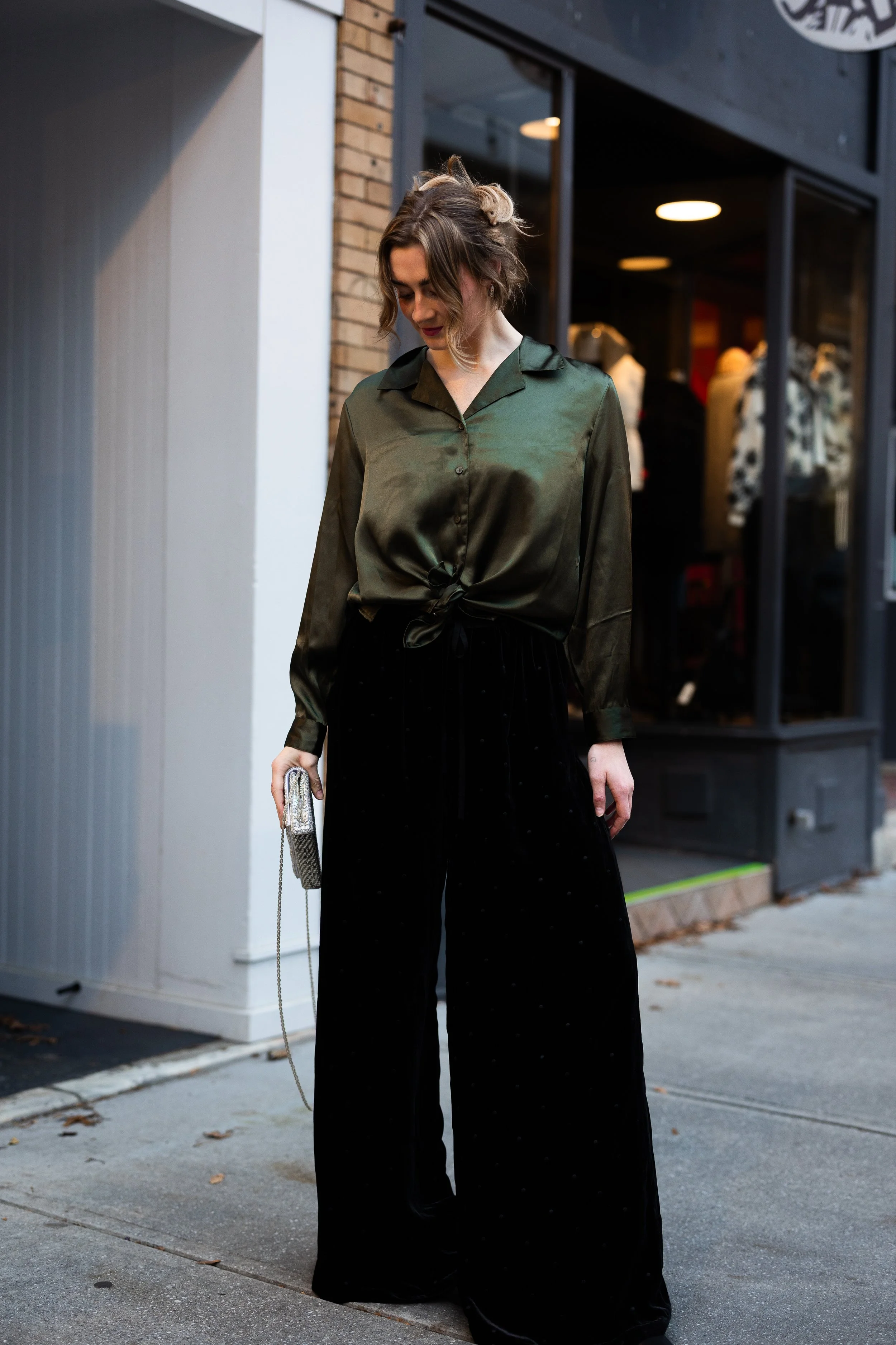 A woman standing on the sidewalk outside a store, wearing a green satin button-up shirt tied at the waist and black wide-leg pants. She is holding a silver clutch with a chain strap.