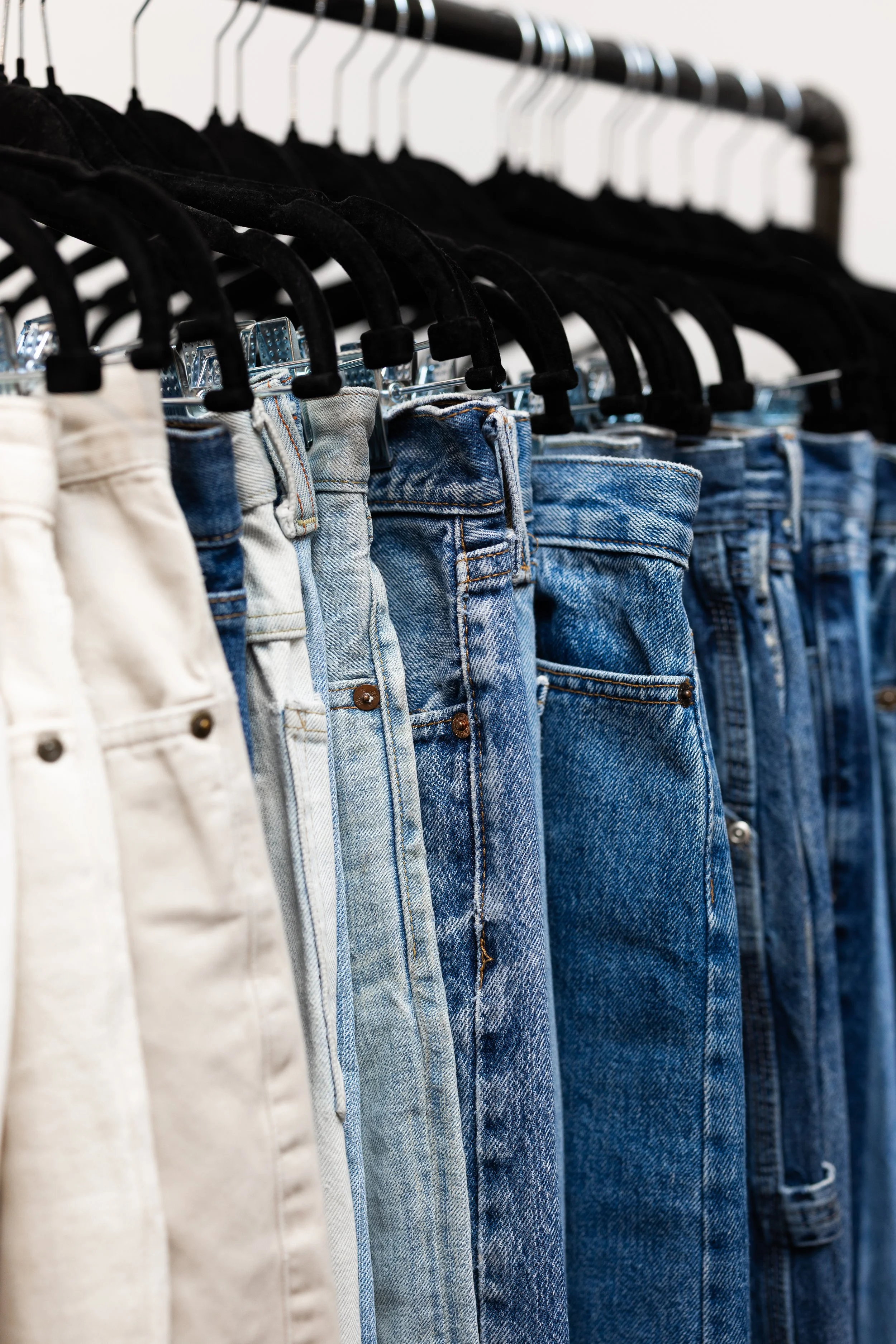 A row of jeans in different shades of blue and white hanging on a clothing rack with black hangers.