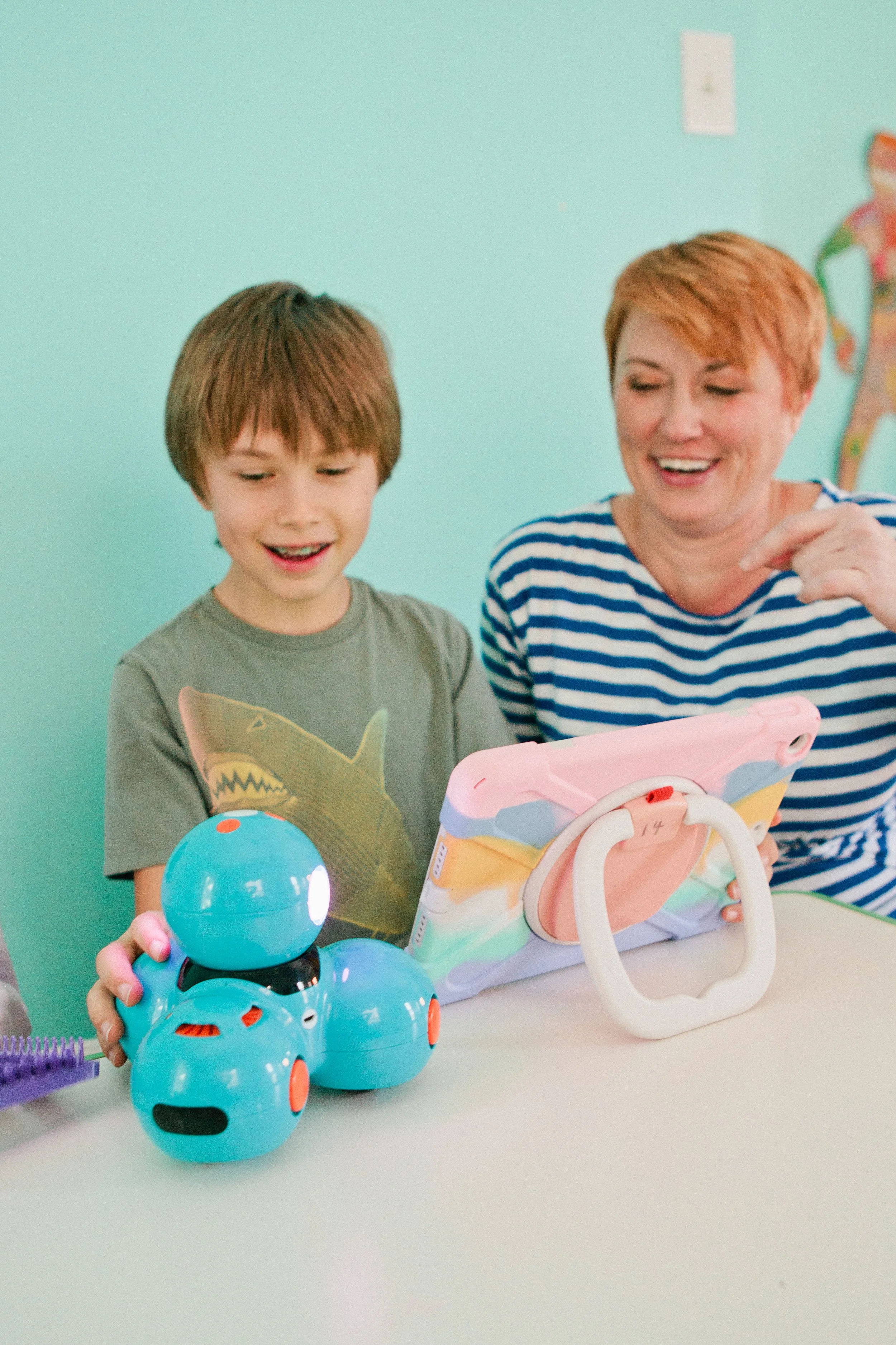 A young boy assisted by his teacher in a colorful room. There are three blue robotic toys on the table, one of which is stacked on top of the others.