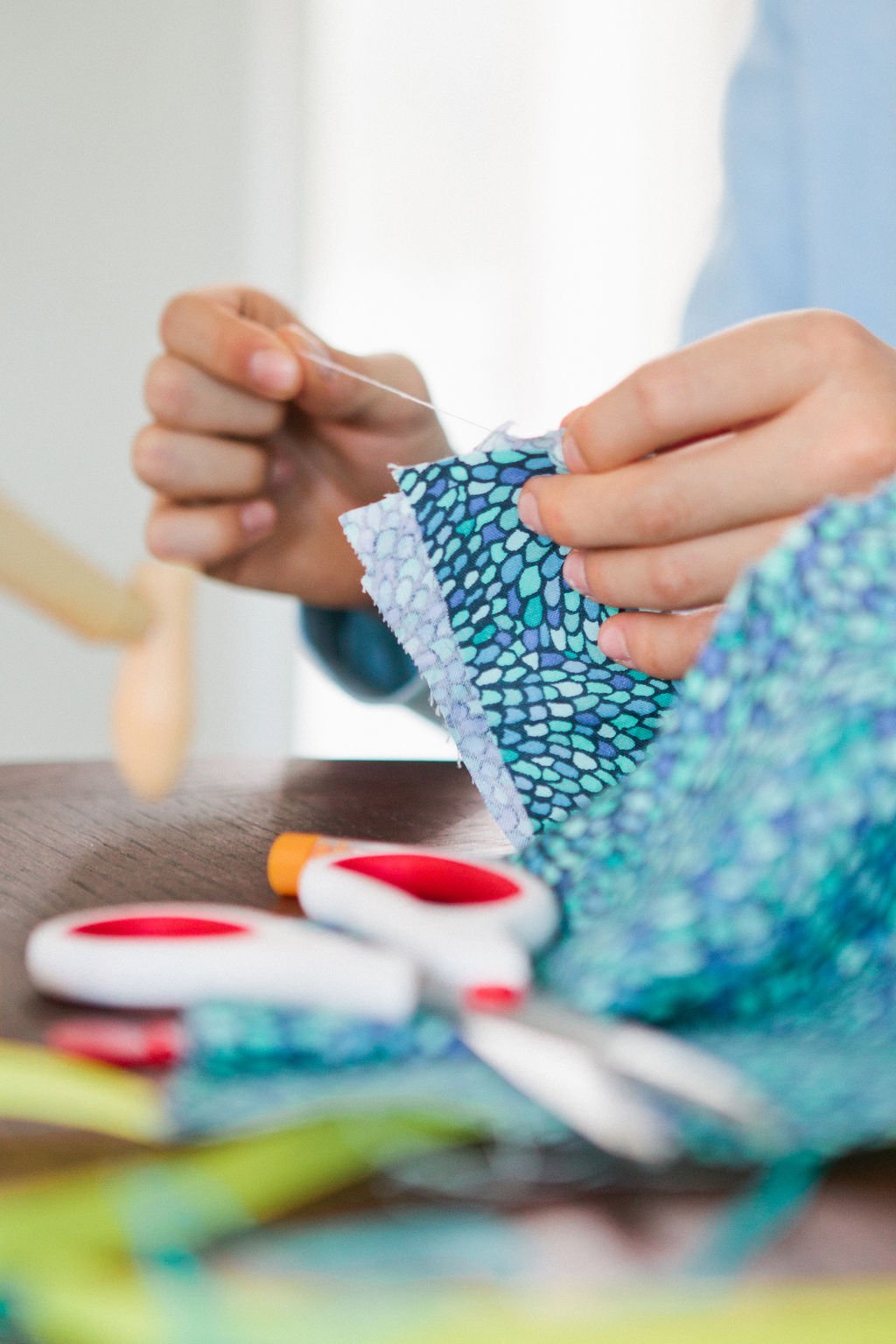 Person sewing fabric with a needle and thread, scissors and sewing supplies on a wooden surface.