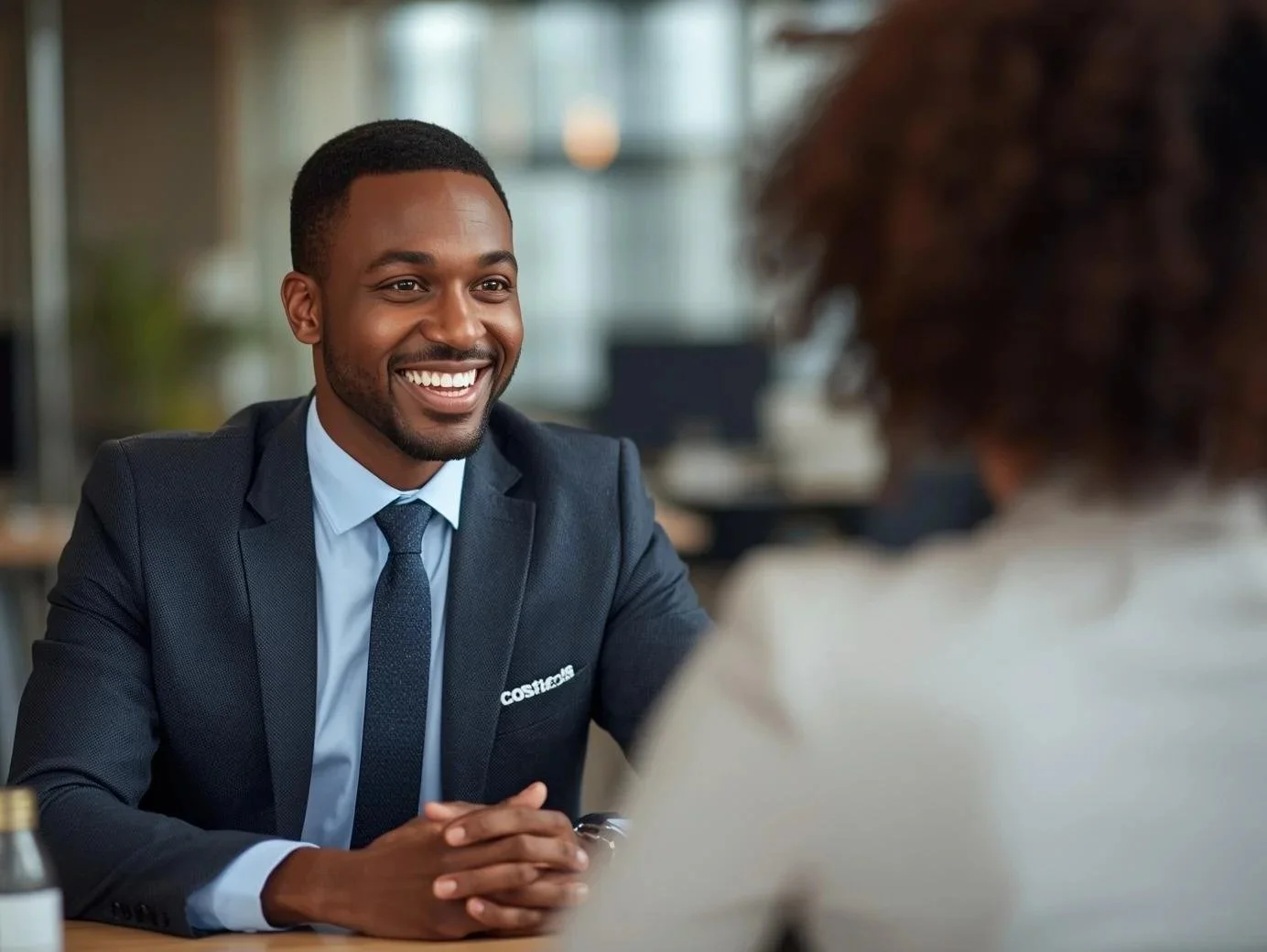 A man in a suit smiling during a interview, talking with a woman in a white shirt.