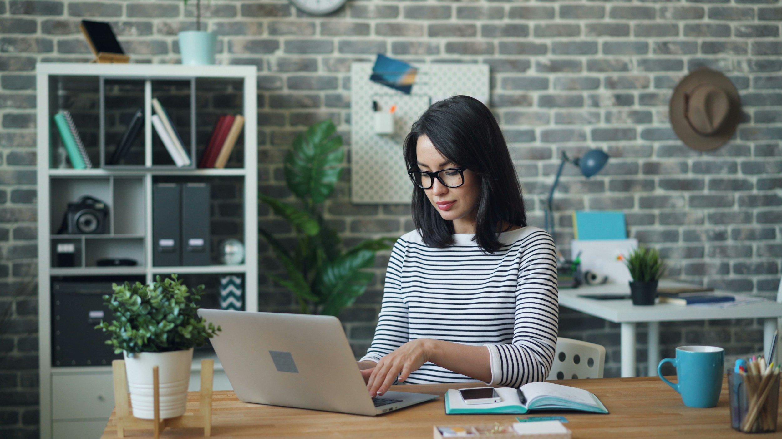 A woman with black hair and glasses working on a laptop at a wooden desk in a modern office with a brick wall. There are plants, a notebook, a mug, and office supplies on the desk, with shelves and a whiteboard in the background.