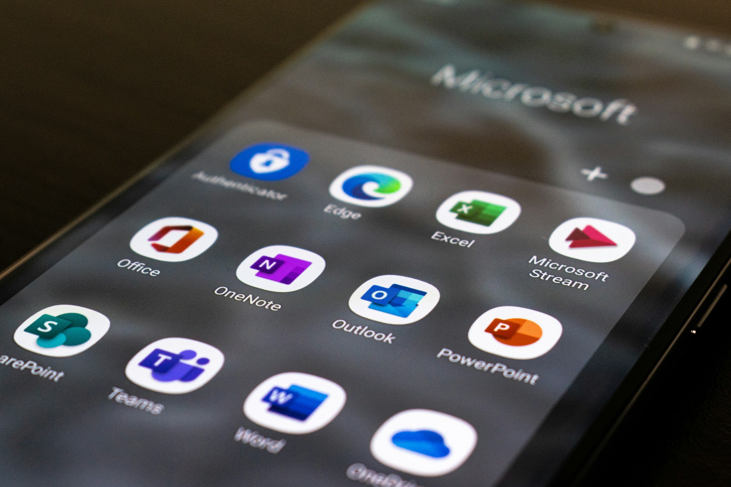 Close-up of a smartphone screen displaying a folder named 'Microsoft' with various app icons including Office, OneNote, Outlook, PowerPoint, Teams, Word, SharePoint, SharePoint, Edge, Microsoft Stream, and other Microsoft apps.