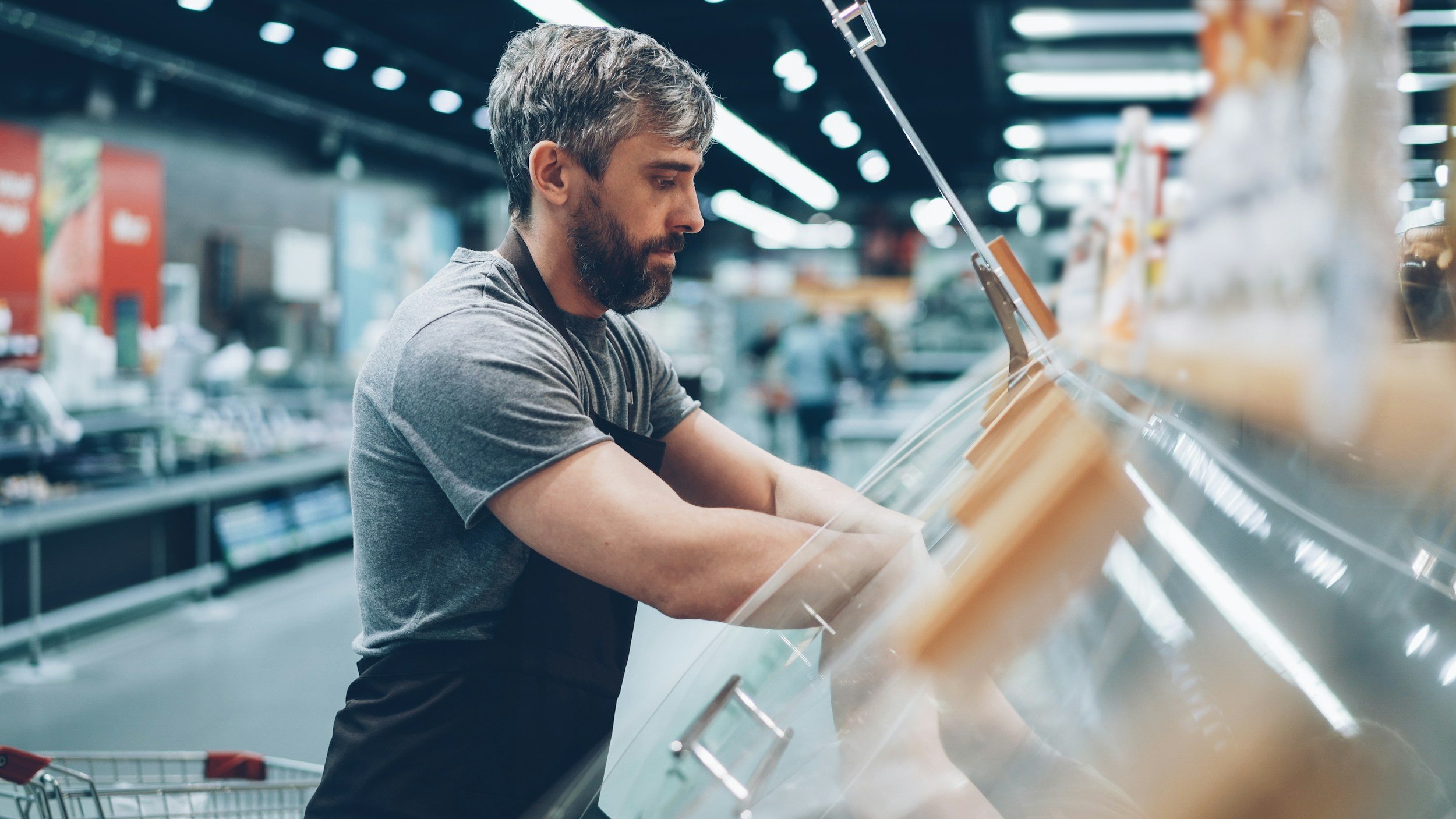 A man with a beard and gray hair shopping at a grocery store, looking into a refrigerated display case.
