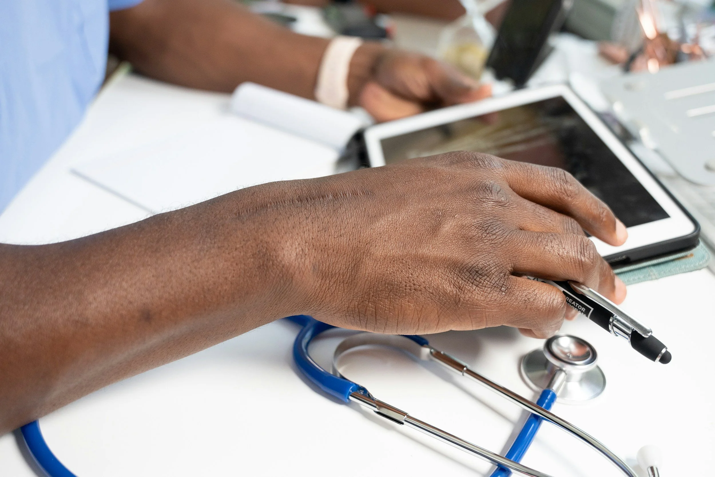 Close-up of a dark-skinned healthcare professional's hand holding a black marker, with a stethoscope and electronic tablets on a white surface, indicating a medical setting.
