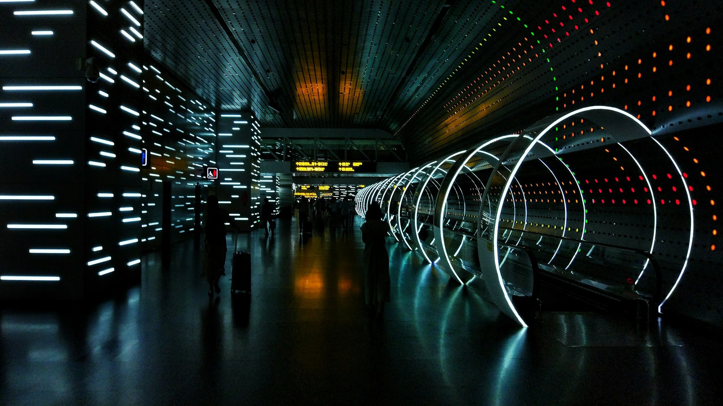Interior of a modern airport terminal with illuminated walls and a futuristic design, featuring circular tunnel-like structures and digital signage.