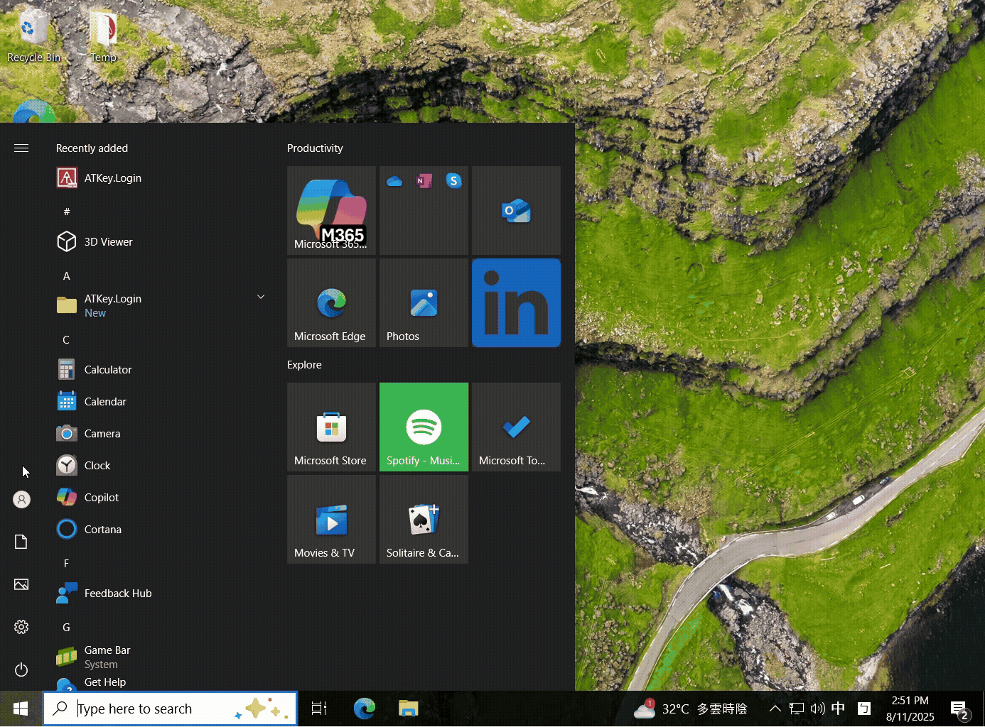 A computer desktop with a start menu open, showing app tiles and folders, over a wallpaper of green cliffs and a winding road.