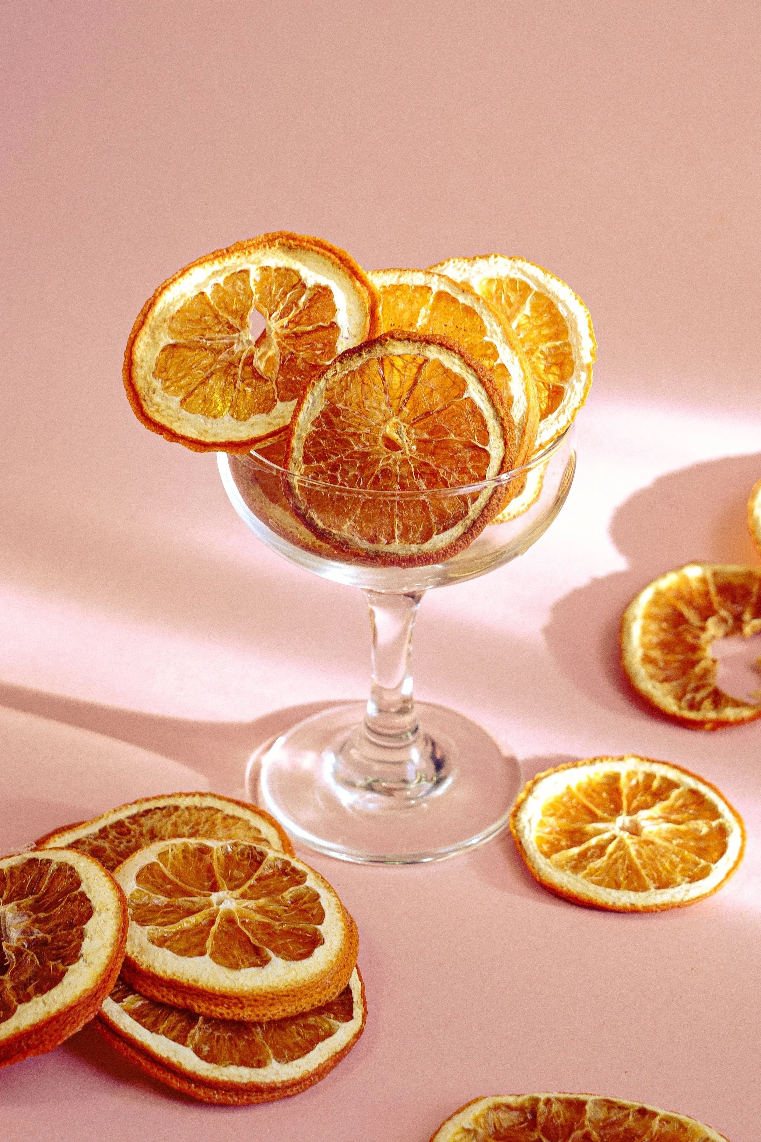 A clear glass coupe filled with dried orange slices, set against a light pink background with some orange slices scattered around the base.