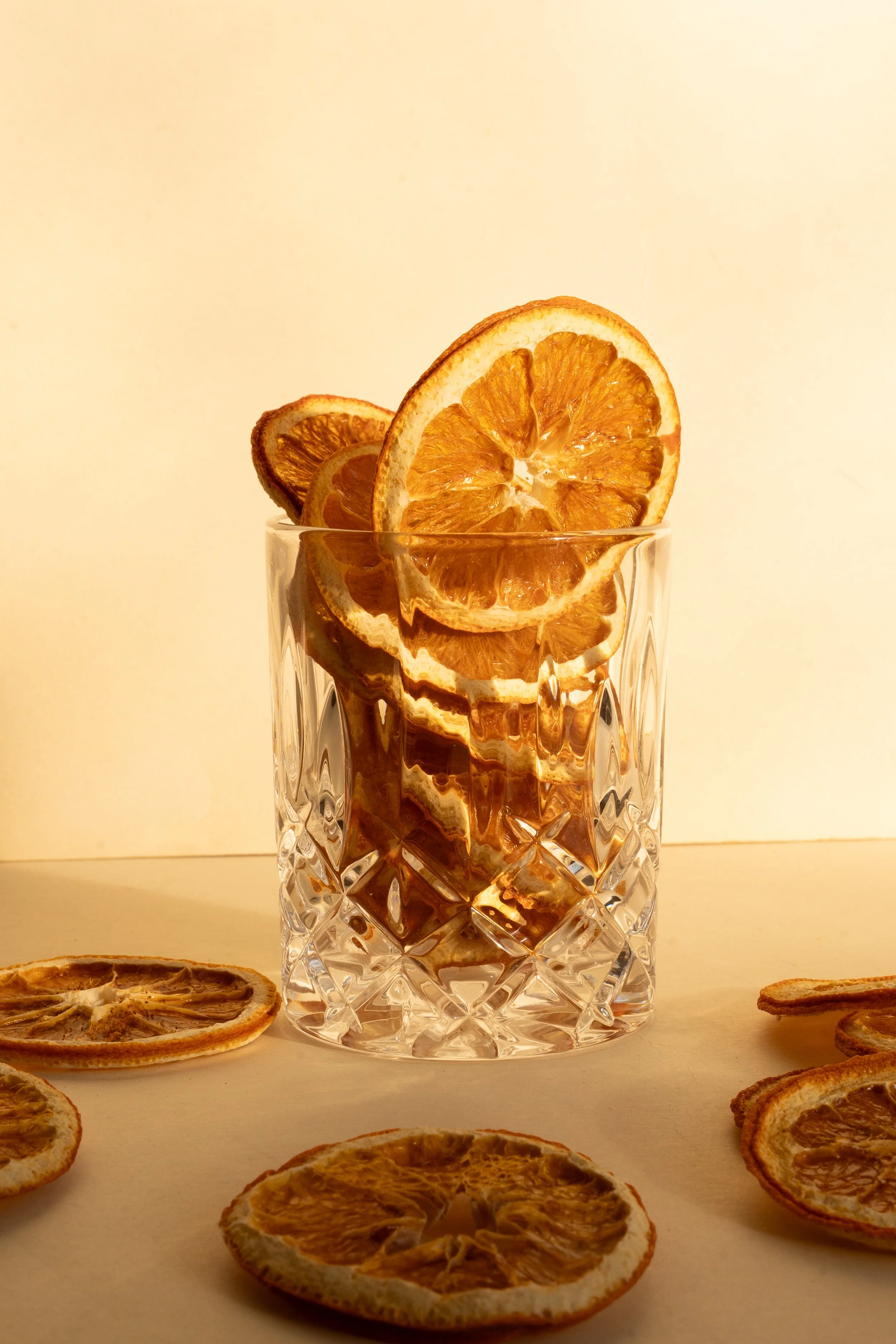 A glass of water with dried orange slices inside and on top, surrounded by additional dried orange slices on a light-colored surface against a plain background.