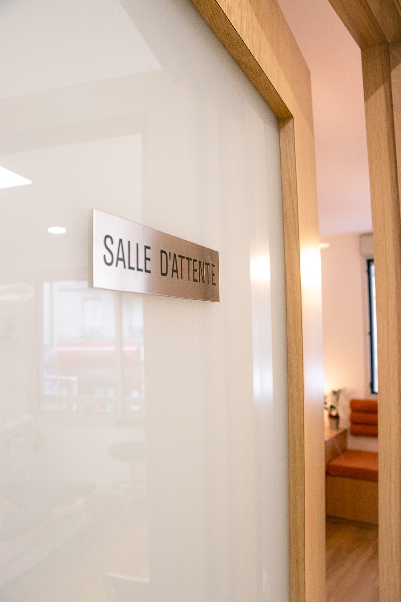 Porte en verre avec une encadrement en bois portant une inscription 'Salle d'attente' en français, donnant sur une salle d'attente avec un canapé et une fenêtre.