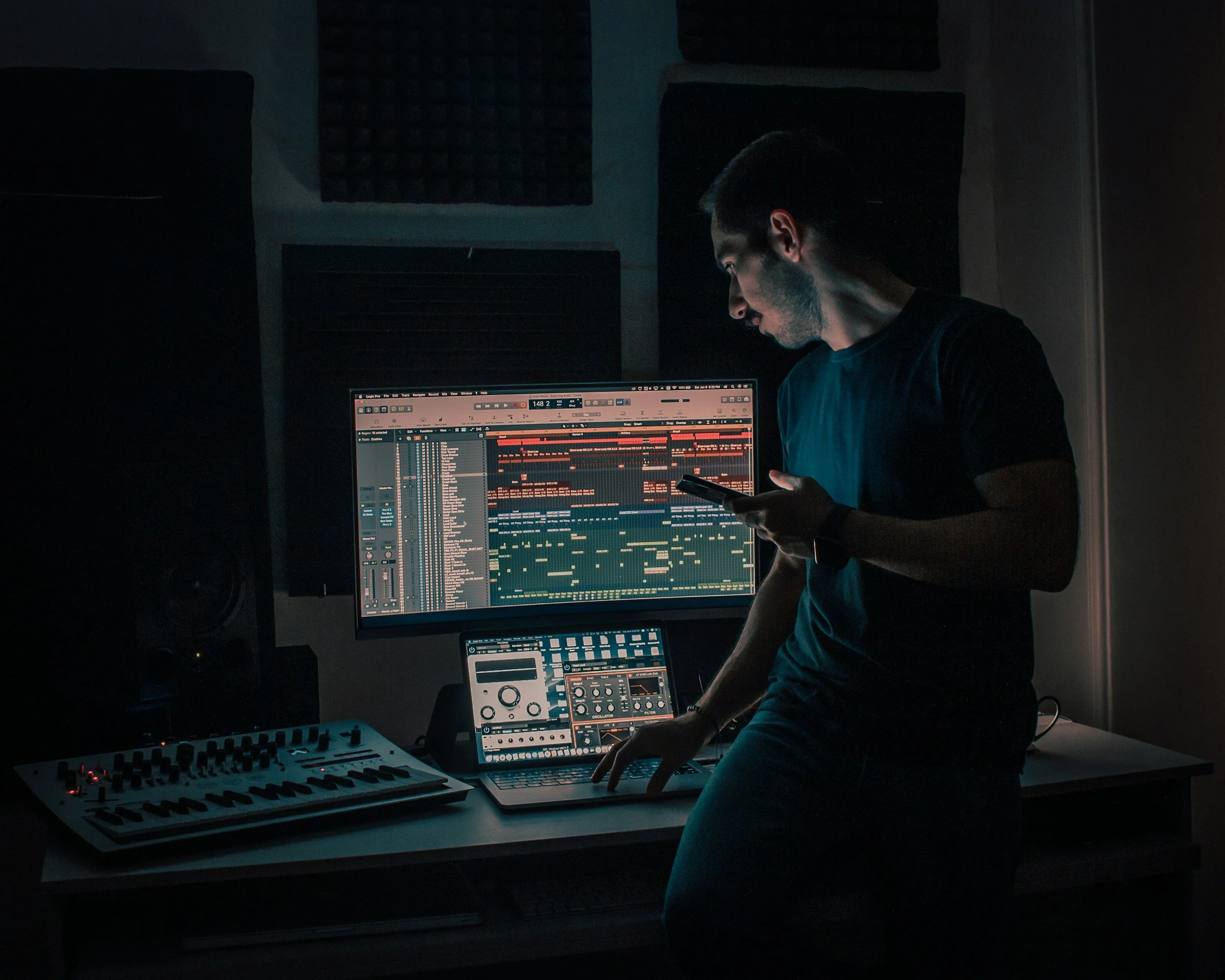 A young man in a dark room, working with music production software on a computer monitor, electronic music equipment, and a keyboard, illuminated by screen light.