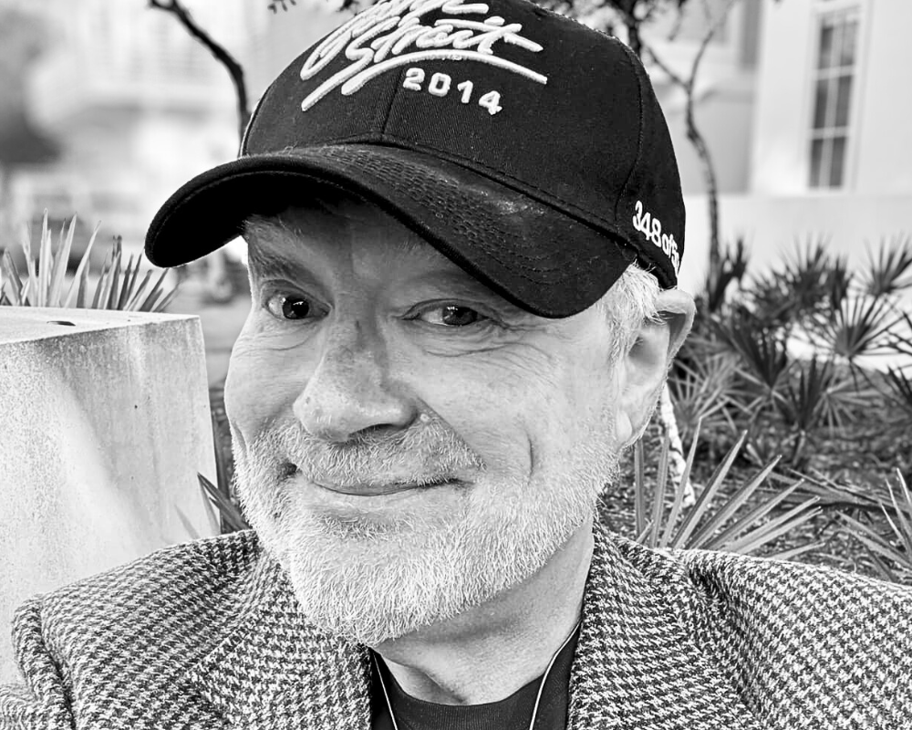 A close-up black and white photo of a smiling man with white hair and beard, wearing a baseball cap and a patterned jacket, outdoors with plants and trees in the background.