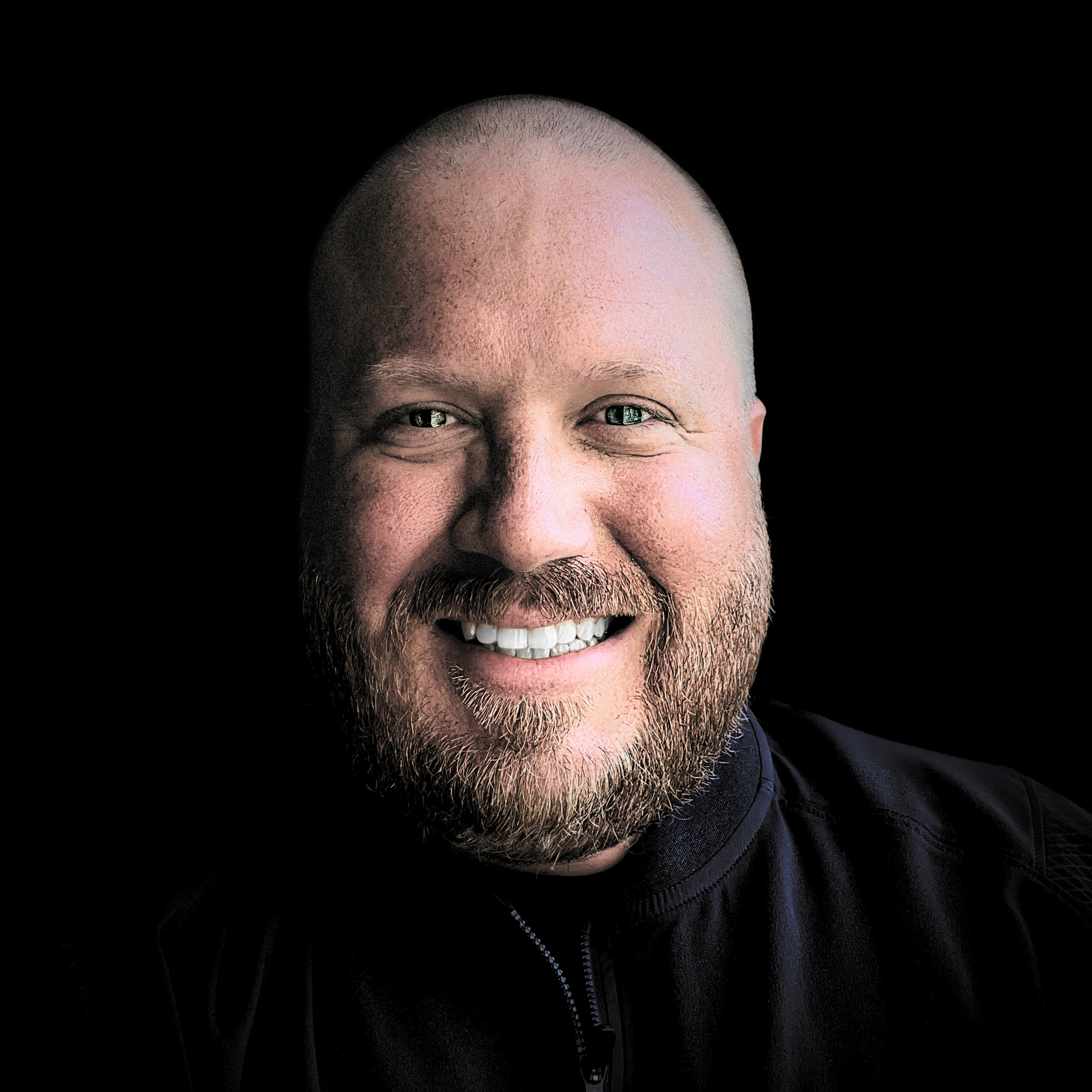 Close-up portrait of a smiling rob coates with a beard, fair skin, and short hair, wearing a black shirt against a black background.