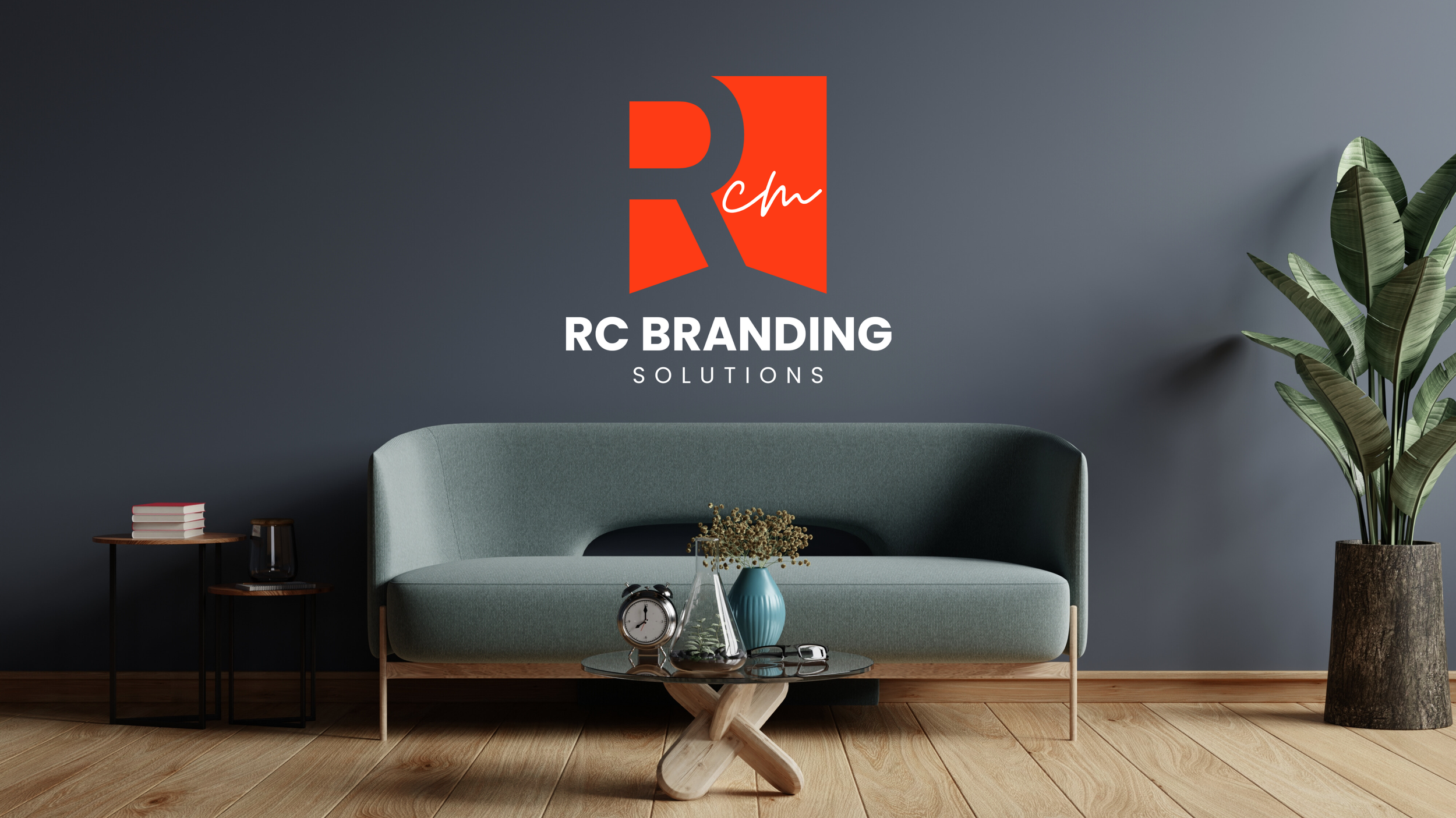 Modern living room with a dark grey wall, a light green sofa, two small black side tables with books and a lantern, a wooden coffee table with vases, a clock, and glasses, a tall potted plant, and a large logo on the wall that reads "RC BRANDING SOLUTIONS."
