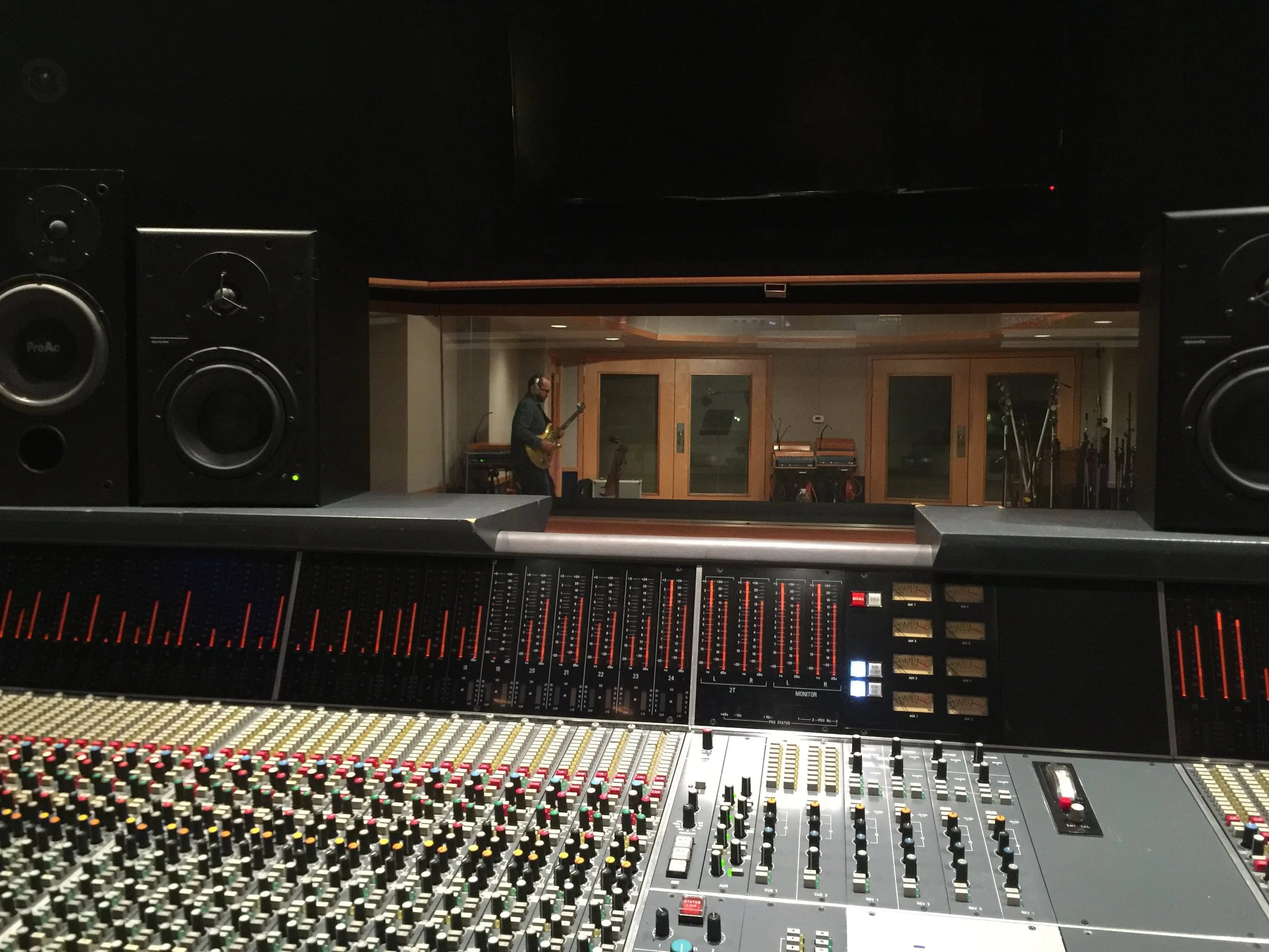 Recording studio view showing a sound mixing console in the foreground, large speakers on either side, and a recording room in the background with a musician playing guitar.