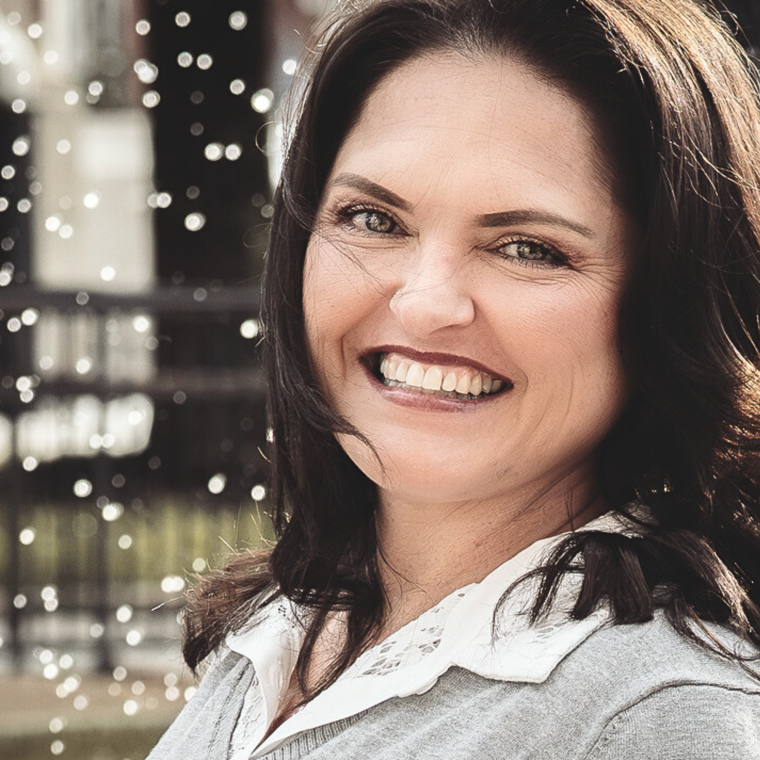 Close-up of a woman with dark hair smiling outdoors with background lights.