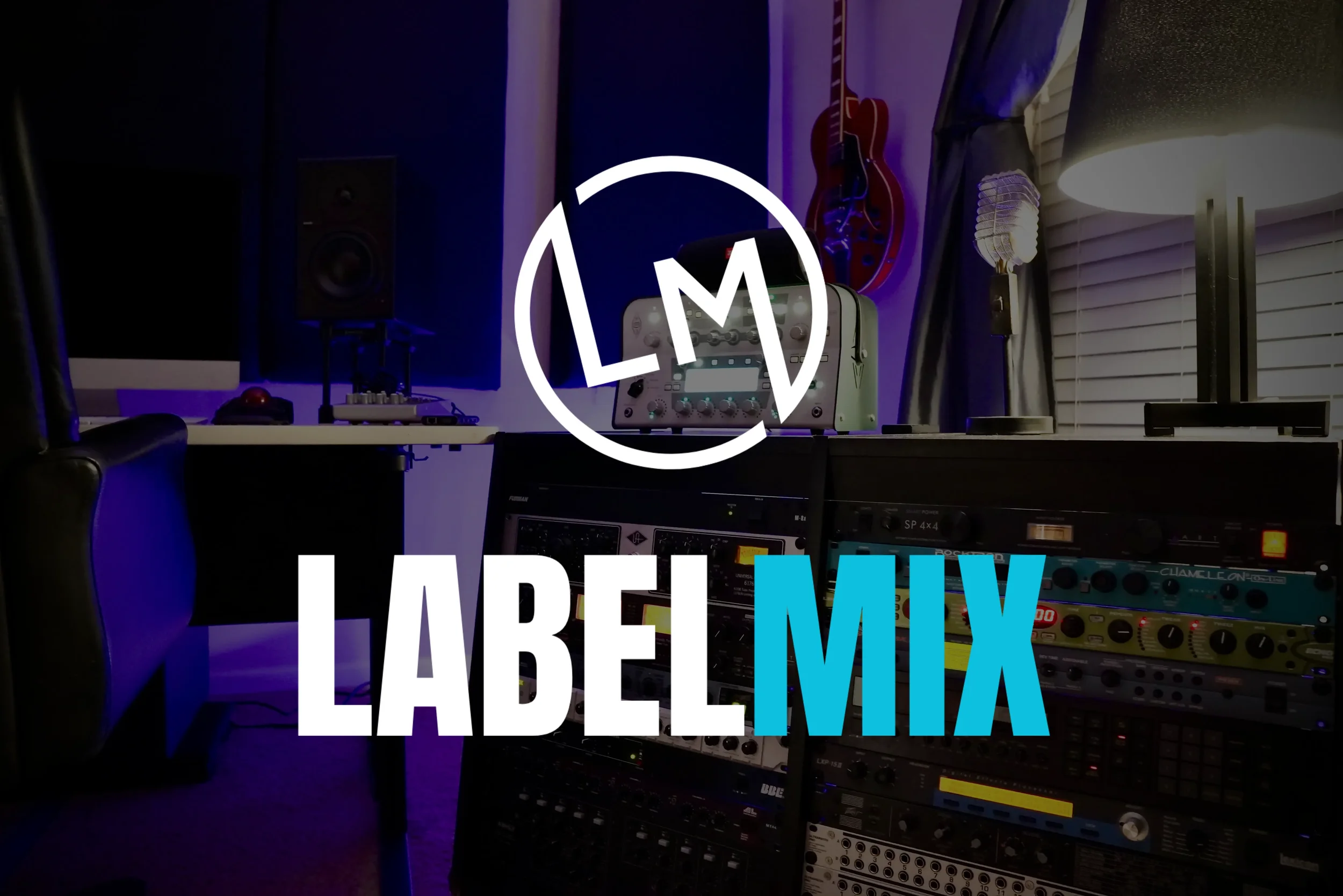 Music studio with mixing equipment, a guitar on the wall, a computer monitor, speakers, and a table lamp in a dimly lit room.