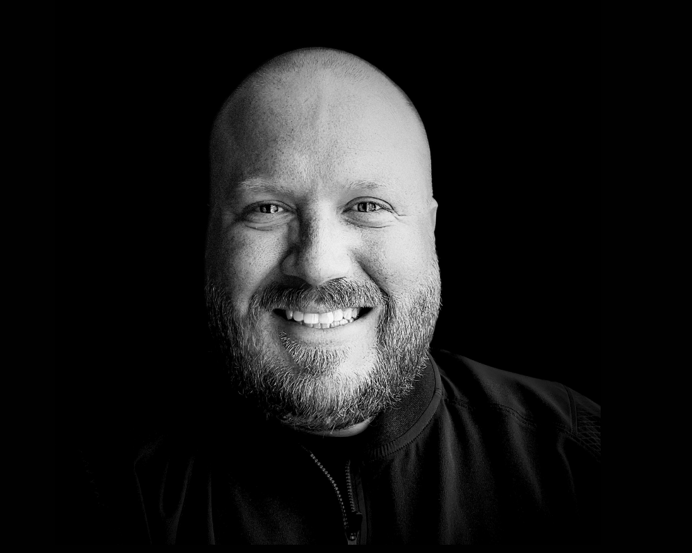 Black and white portrait of a smiling man with a beard and short hair, wearing a dark jacket, against a plain dark background.