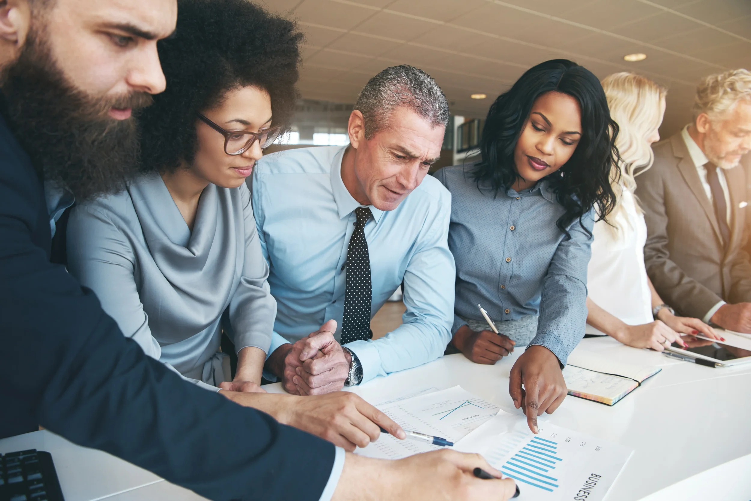 A diverse group of professionals, including men and women of different ethnicities, engaged in a discussion around a table with documents and charts.